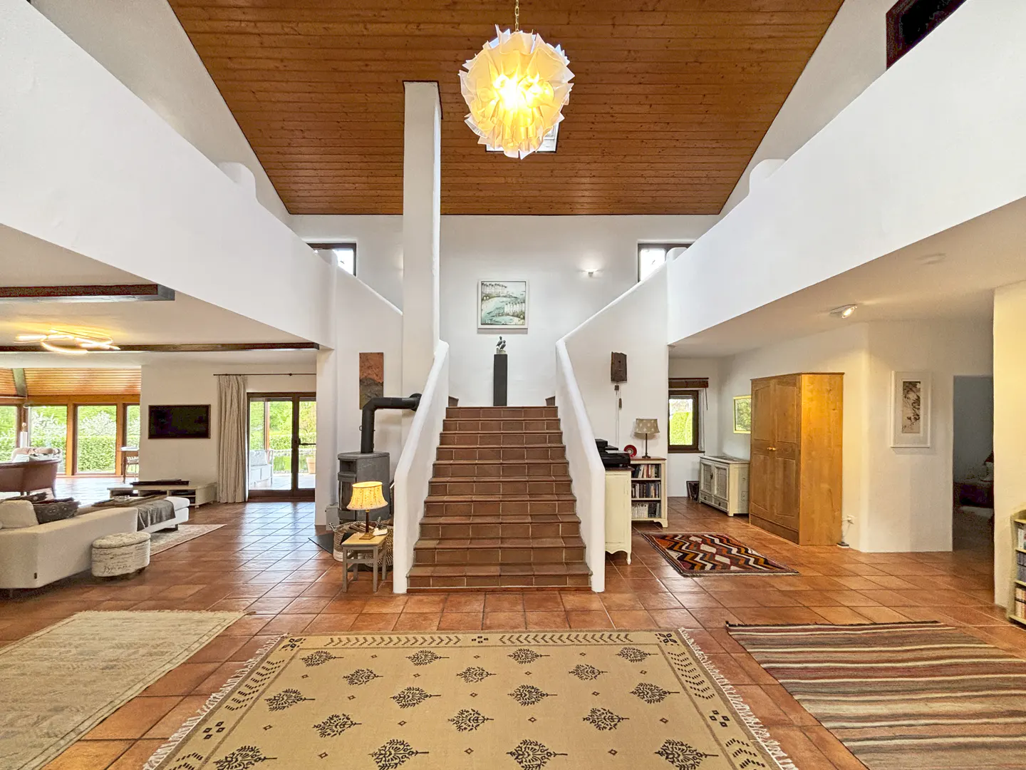 Bright foyer with terracotta tile floor, white walls, and wood ceiling. Staircase leads to upper level. Rugs and furniture add warmth.