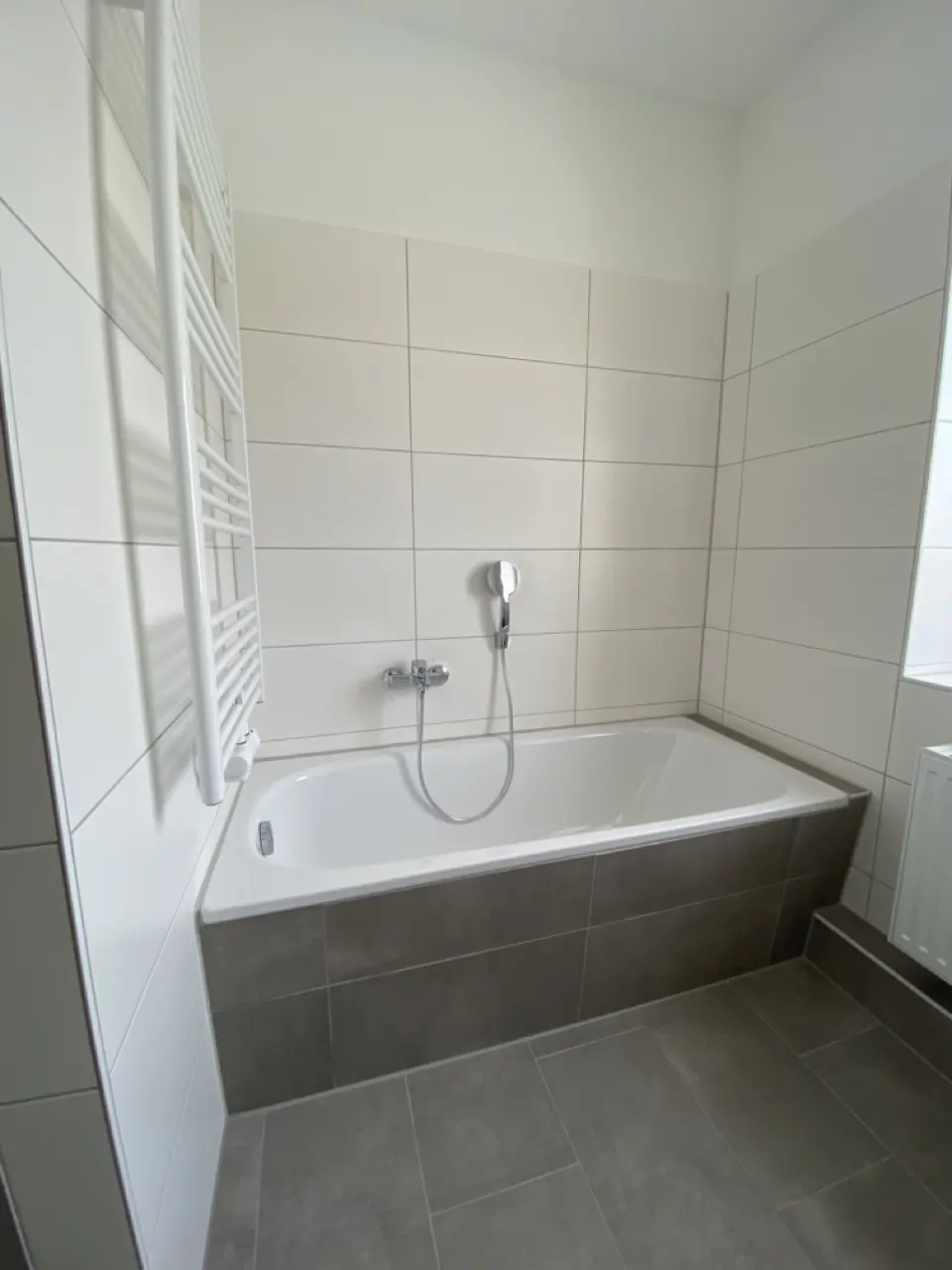 Modern bathroom with a white bathtub, grey tiled base, white wall tiles, and a white towel radiator.