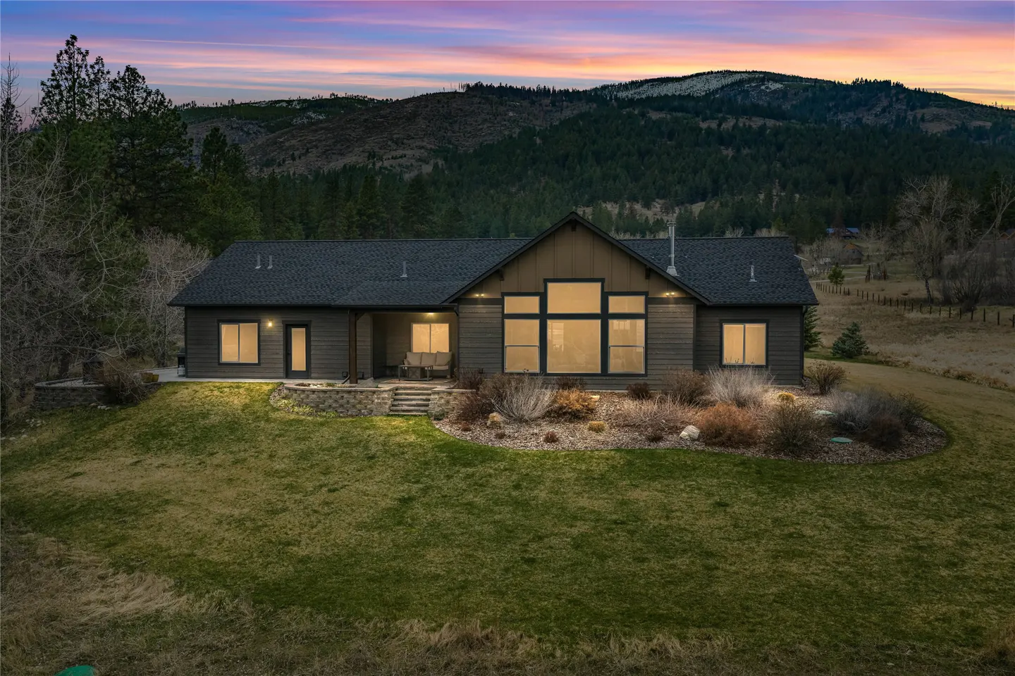 A single-story home with a dark roof and siding sits on a green lawn with mountains in the background.