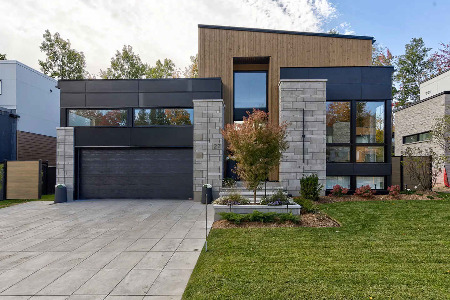 Modern two-story home with a black garage door, stone pillars, wood siding, and a green lawn.