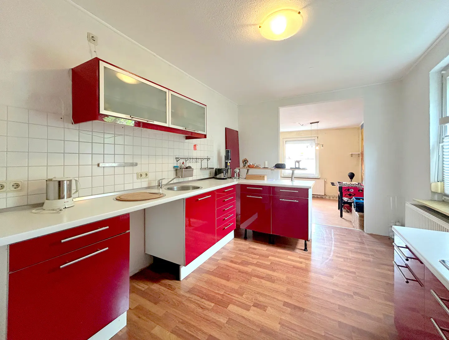 A bright kitchen with red cabinets, white countertops, and wood floors. A doorway leads to another room.