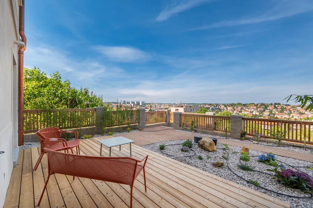 Wooden deck with red chairs, table, and rock garden overlooking a city under a blue sky.
