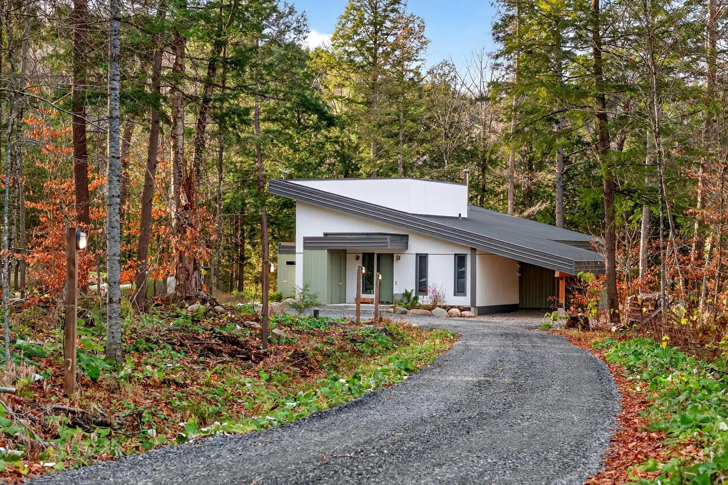 Modern white house with a gray roof, nestled in a forest with a gravel driveway leading up to it.