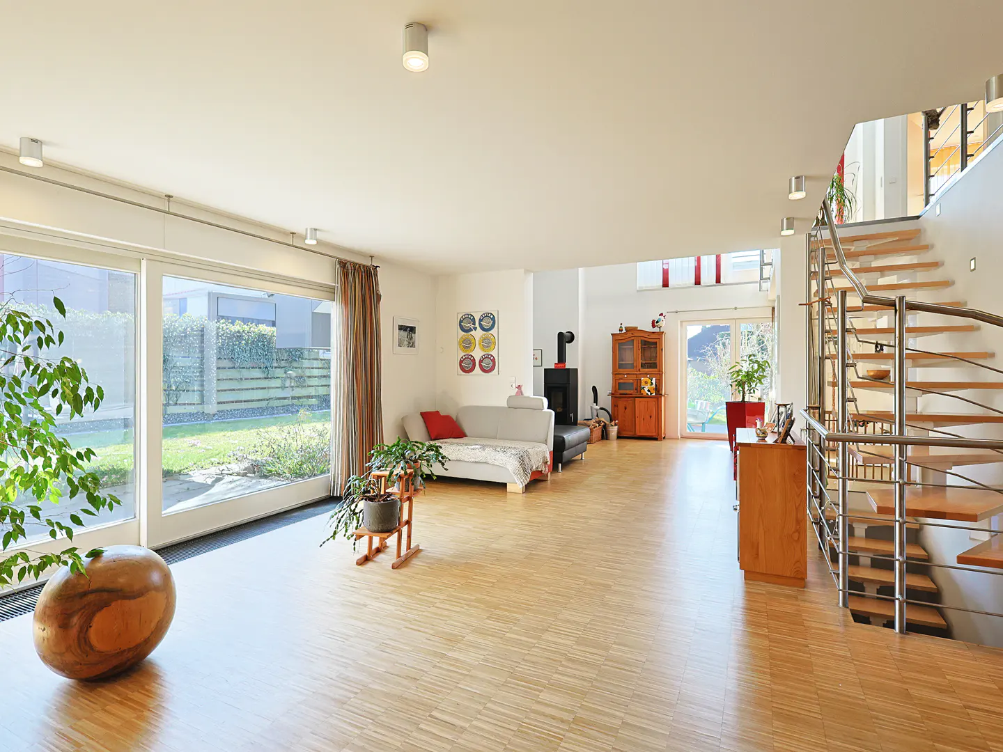 Bright, open living room with wood floors, large windows, and a spiral staircase. A gray sofa and wood-burning stove add warmth.