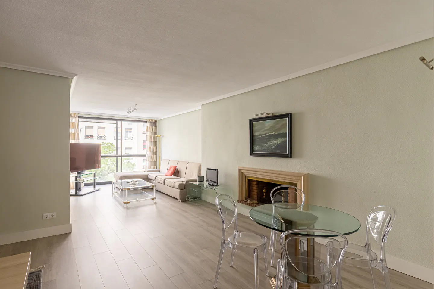 Bright living room with light wood floors, a beige sectional sofa, glass dining table, and a fireplace with wall art.