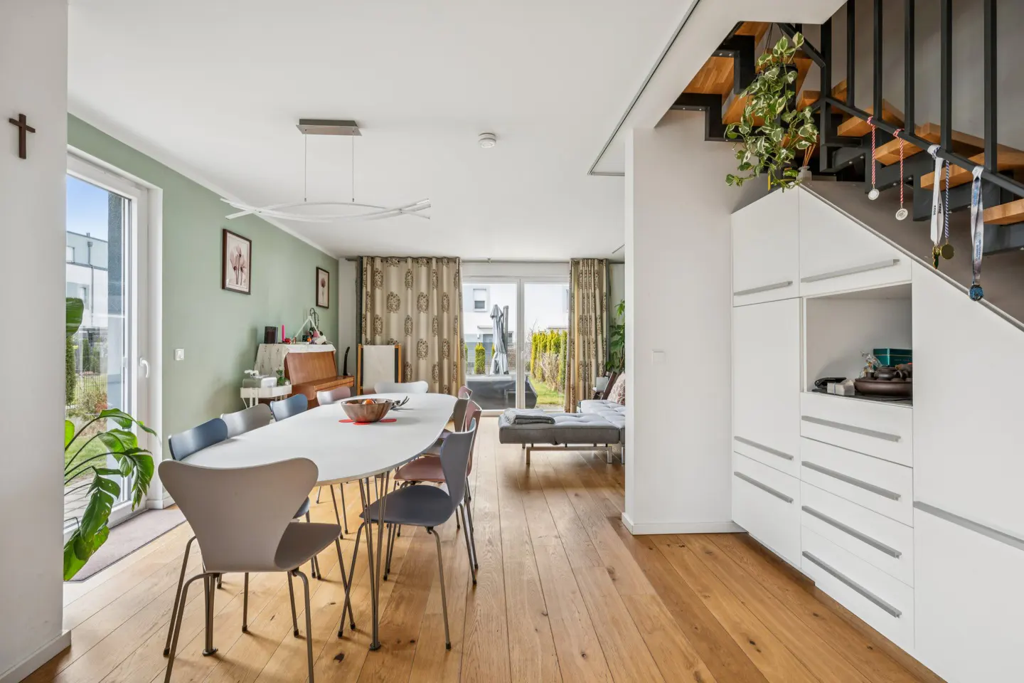 Modern dining area with white oval table, colorful chairs, hardwood floors, and built-in storage under a wooden staircase.