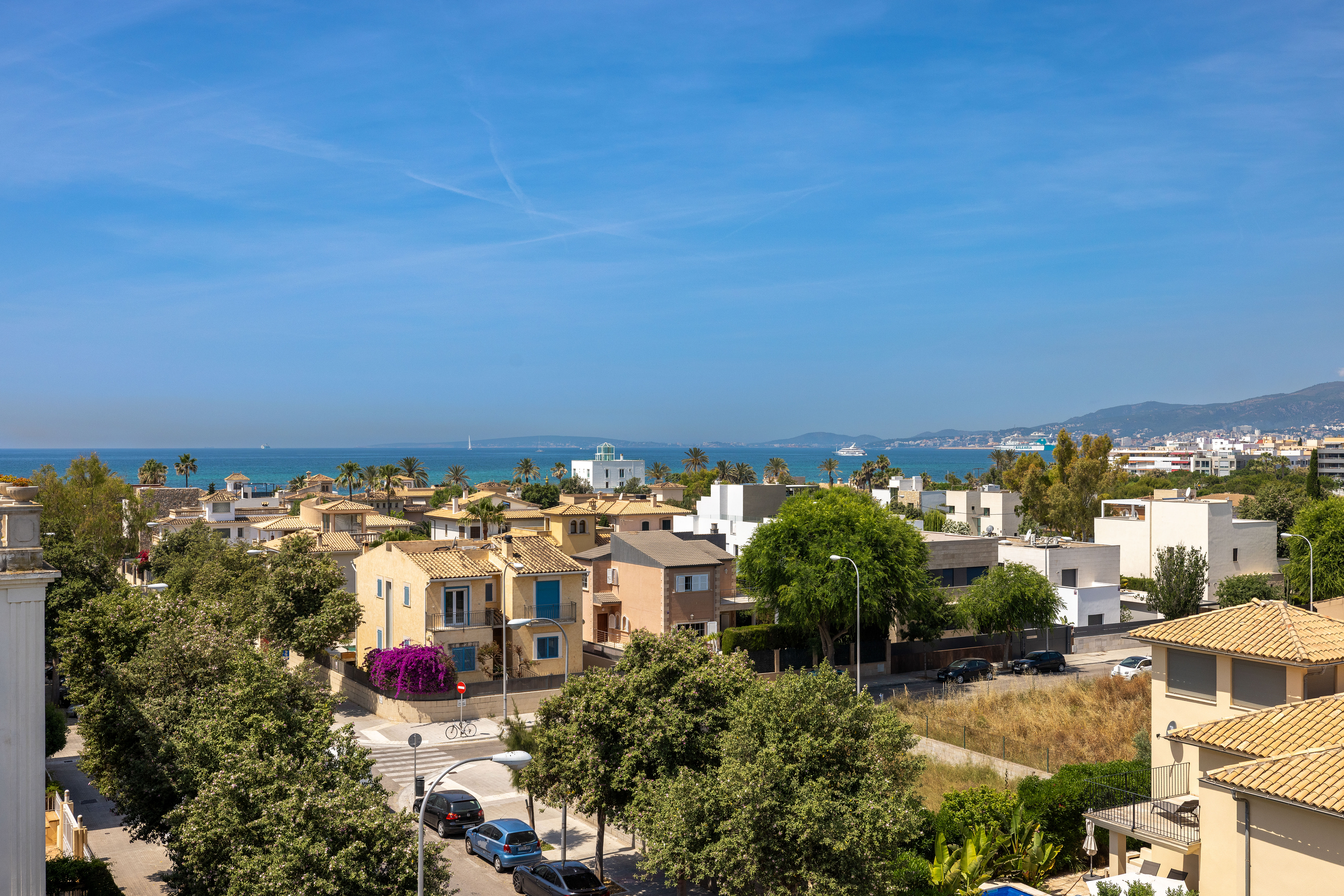 Elevated view of a coastal neighborhood with Mediterranean-style houses, lush green trees, and the blue sea in the distance. [AI-generated text]