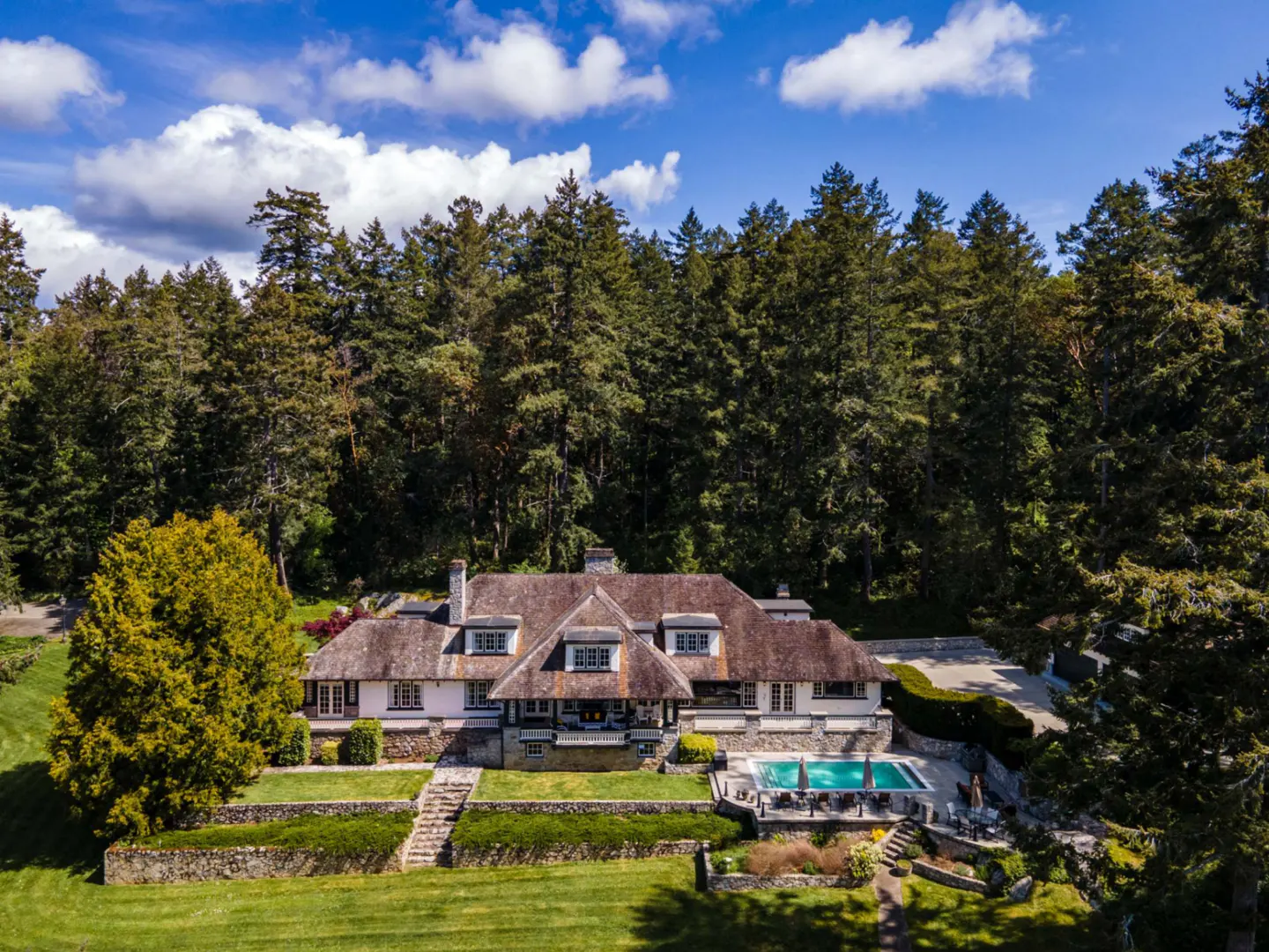Aerial view of a large, white house with a pool, surrounded by green lawn and tall trees under a blue sky.
