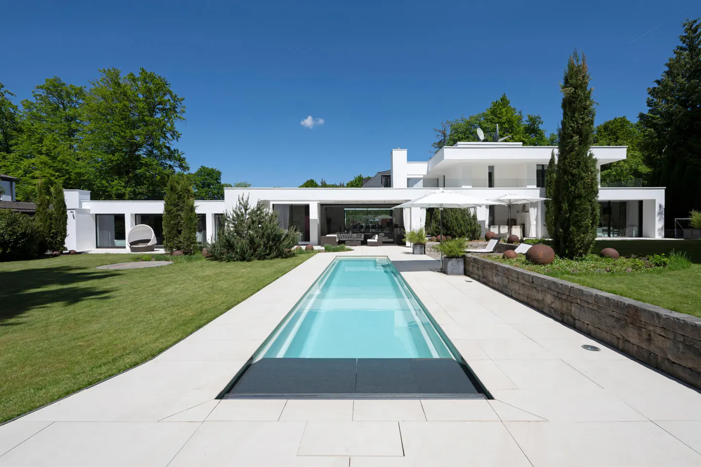 Modern white house with a long, narrow pool. The pool has a retractable cover. Green lawn and trees surround the house. Blue sky.