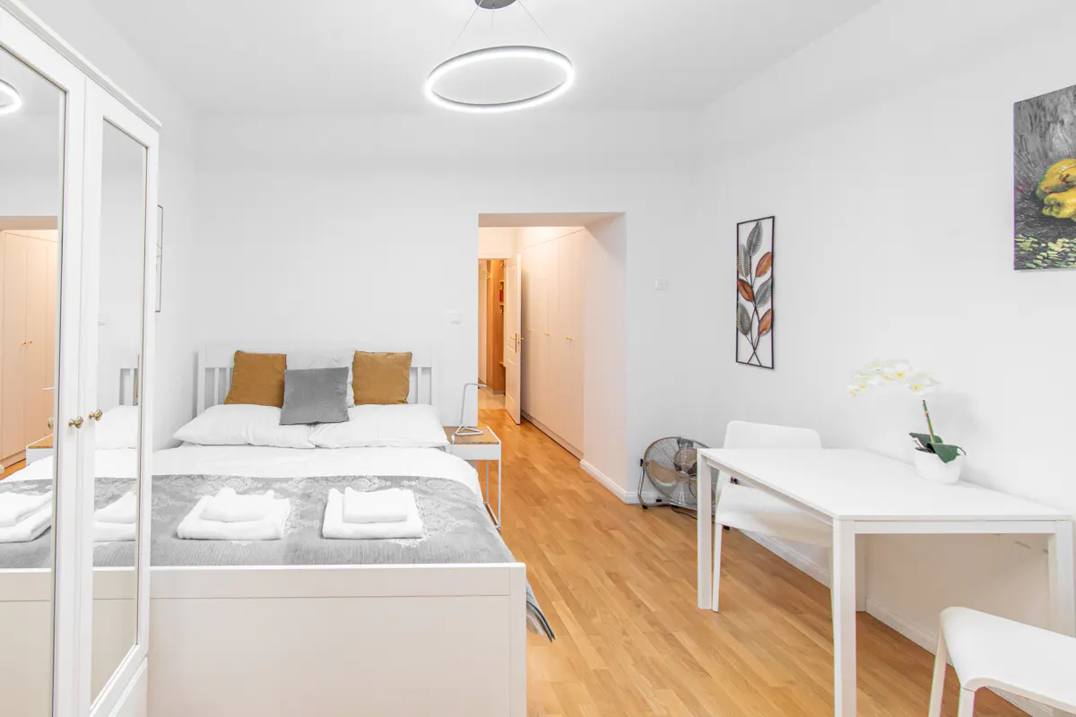 Bright studio apartment with white walls, wood floors, and a bed with yellow and gray pillows. A white table and chairs sit near a fan.