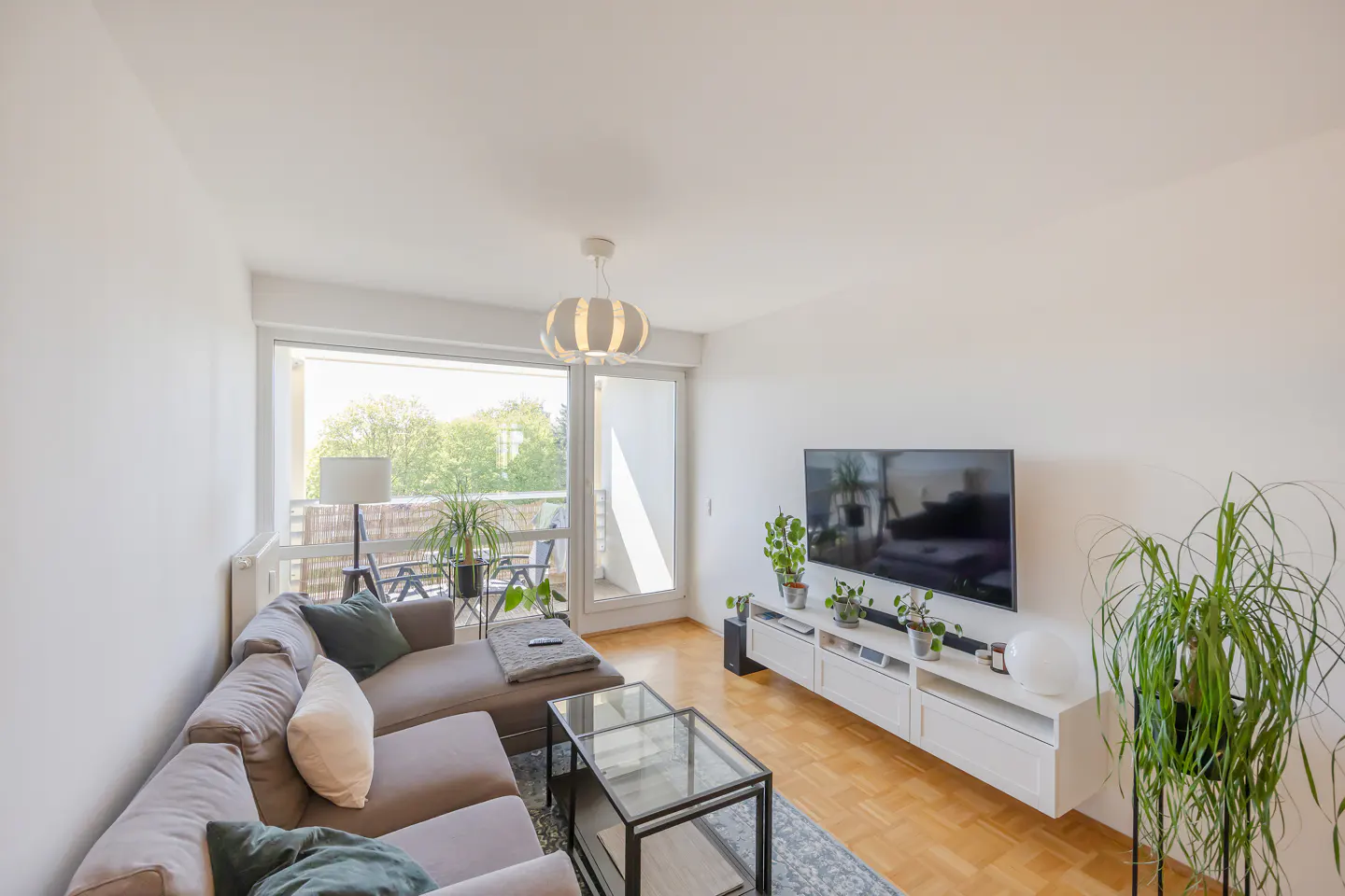 Living room with a gray sectional sofa, glass coffee table, TV, and balcony with chairs. The walls are white, and the floor is wood.