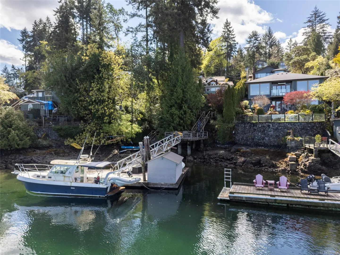 Waterfront property with a boat docked. A modern house sits on a hill surrounded by trees. Docks with chairs extend into the water.