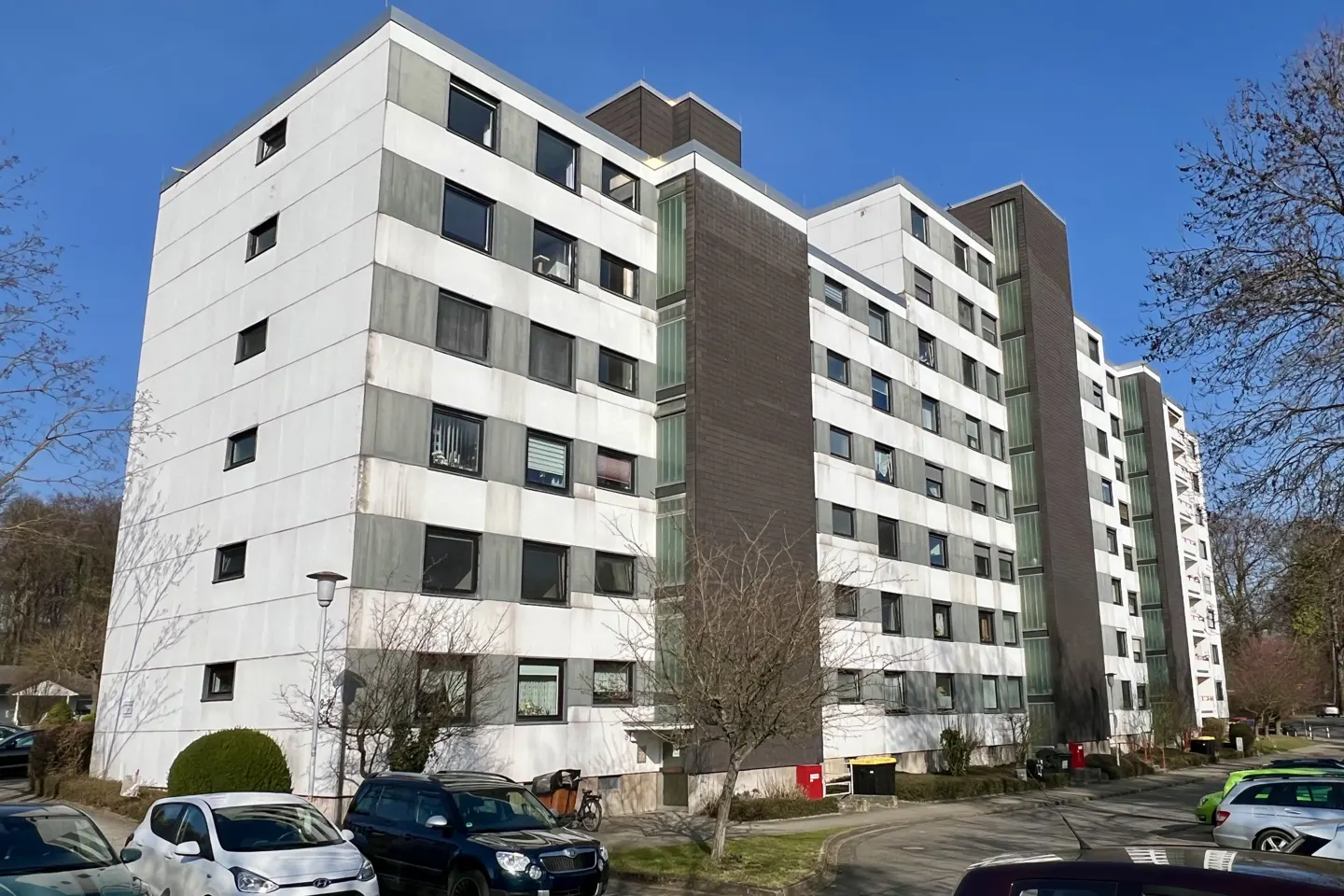Exterior view of a tall, white apartment building with dark gray accents and many windows, surrounded by trees and parked cars.