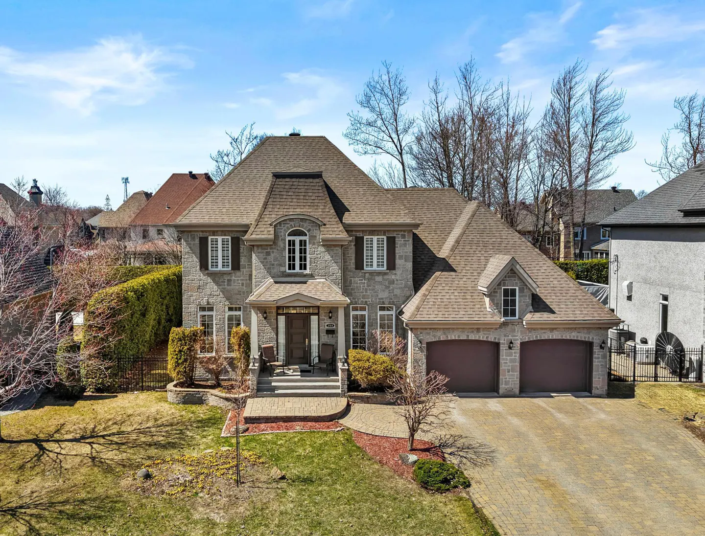 Two-story stone house with a brown roof, two-car garage, and a brick driveway on a sunny day.