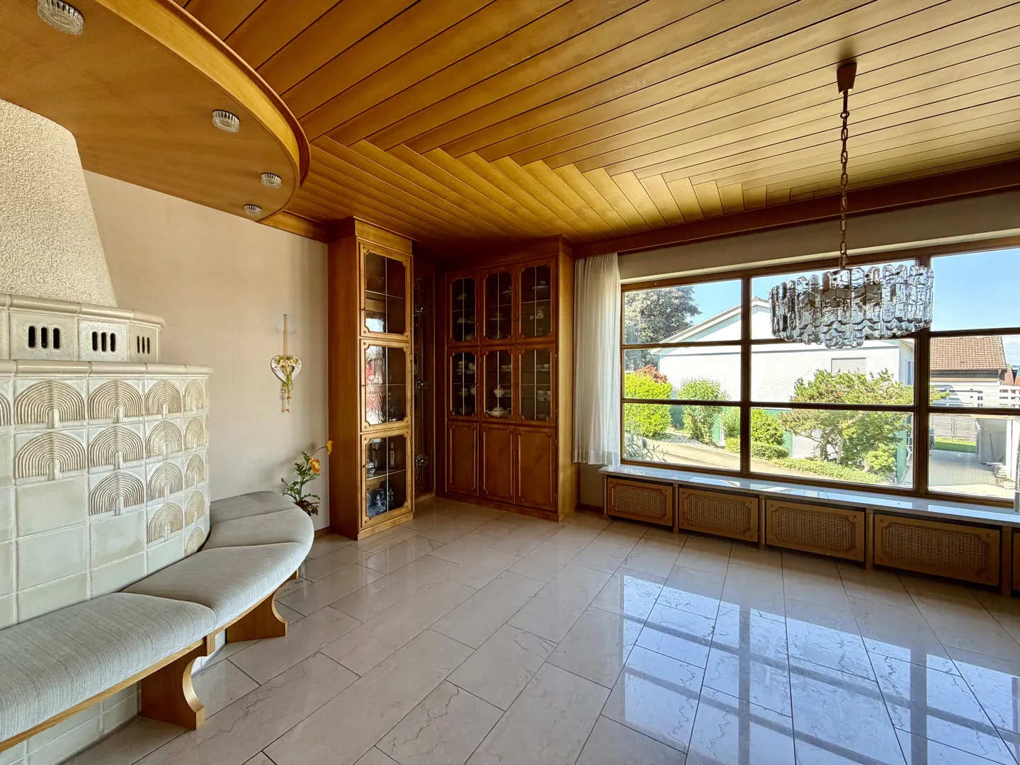 A bright living room with wood paneled ceiling, large window, tiled fireplace with bench, and built-in wooden cabinets.