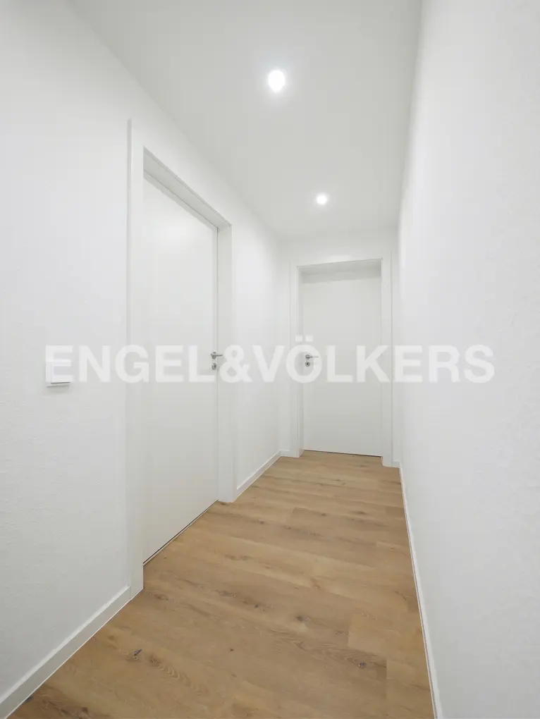 A bright, minimalist hallway with white walls, recessed ceiling lights, and light wood flooring leading to two white doors.