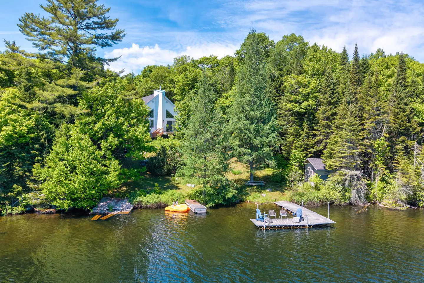 A white A-frame house nestled in a lush green forest by a lake, featuring a private dock with chairs and a yellow kayak.