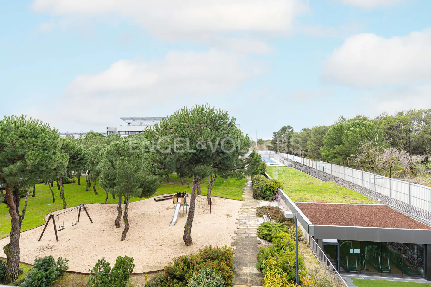View of a playground with swings and a slide, surrounded by trees and greenery, under a cloudy sky.