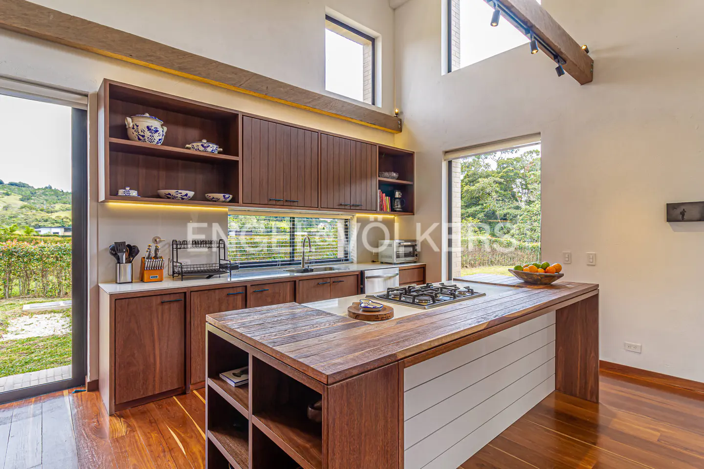 A modern kitchen with wood cabinets, a large island with a gas cooktop, and a view of lush greenery through a large window.