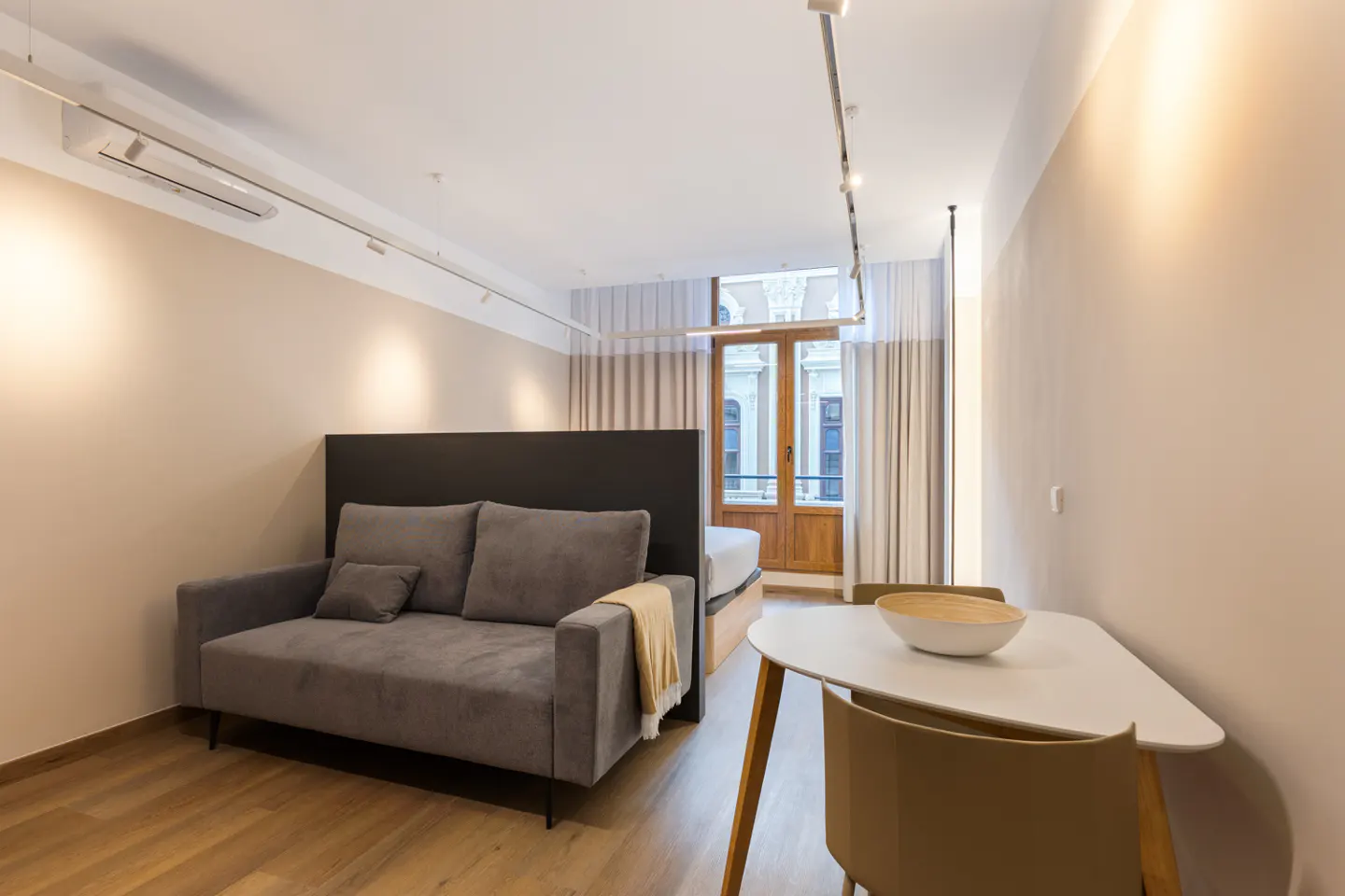 Studio apartment with gray sofa, white table, and wooden floors. A large window with curtains is in the background.