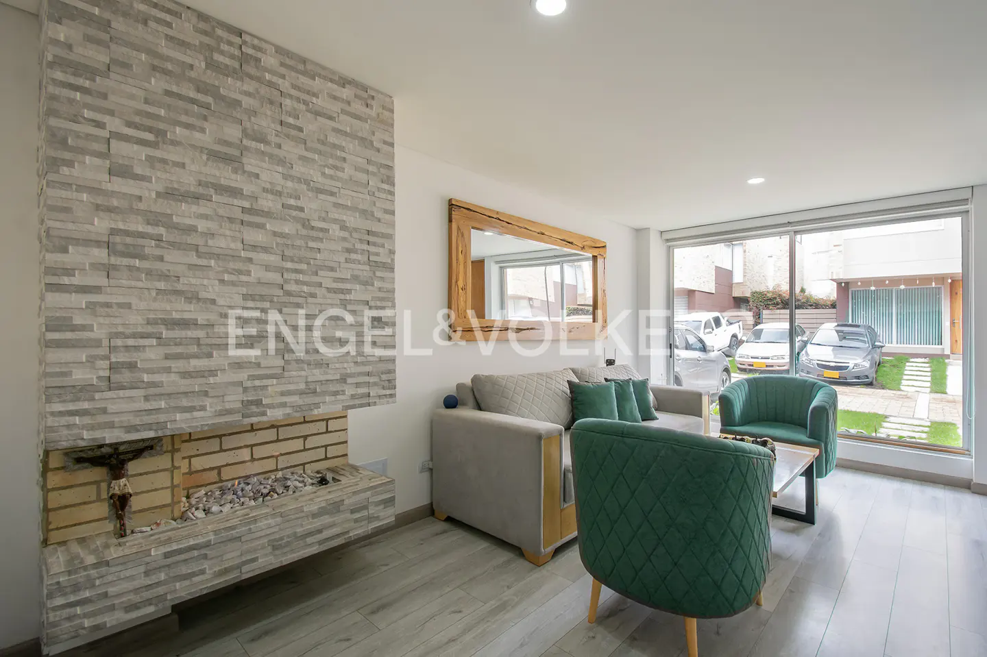 Living room with gray stone fireplace, gray sofa with green pillows, and green armchairs. Large window with view of cars outside.