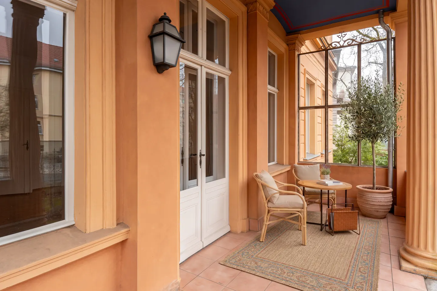 A peach-colored porch with white trim features a table, chairs, and potted tree on a patterned rug.