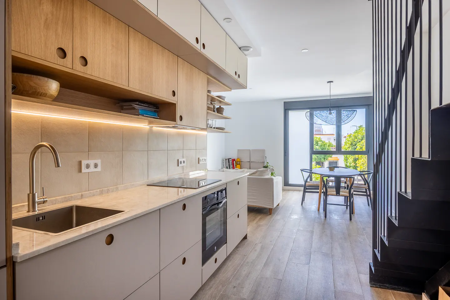 A modern kitchen with light wood cabinets, a stainless steel sink, and a black oven. A dining table and chairs are visible in the background.