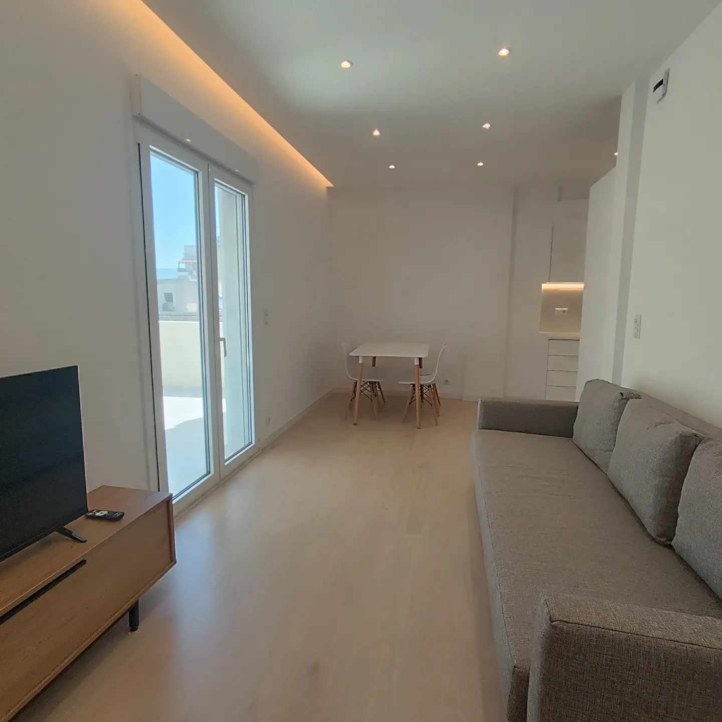 Bright, modern living room with a gray sofa, TV, and a white table with chairs. Sliding glass doors lead to a balcony.