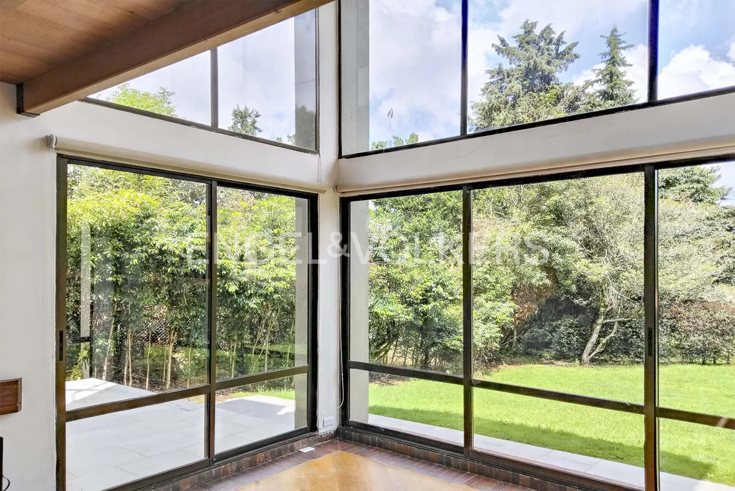 Bright room with large windows overlooking a green garden. The room has wood floors and a wood ceiling beam.