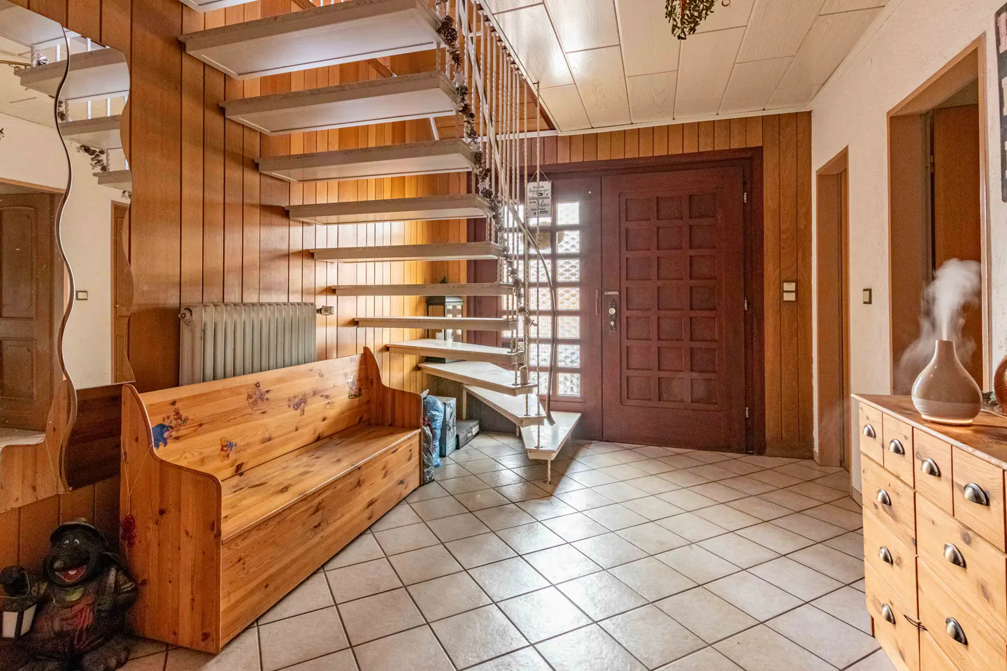 Foyer with wood-paneled walls, tile floor, and a wooden bench. A staircase leads up, and a brown front door is visible. A dresser with a diffuser sits to the right.
