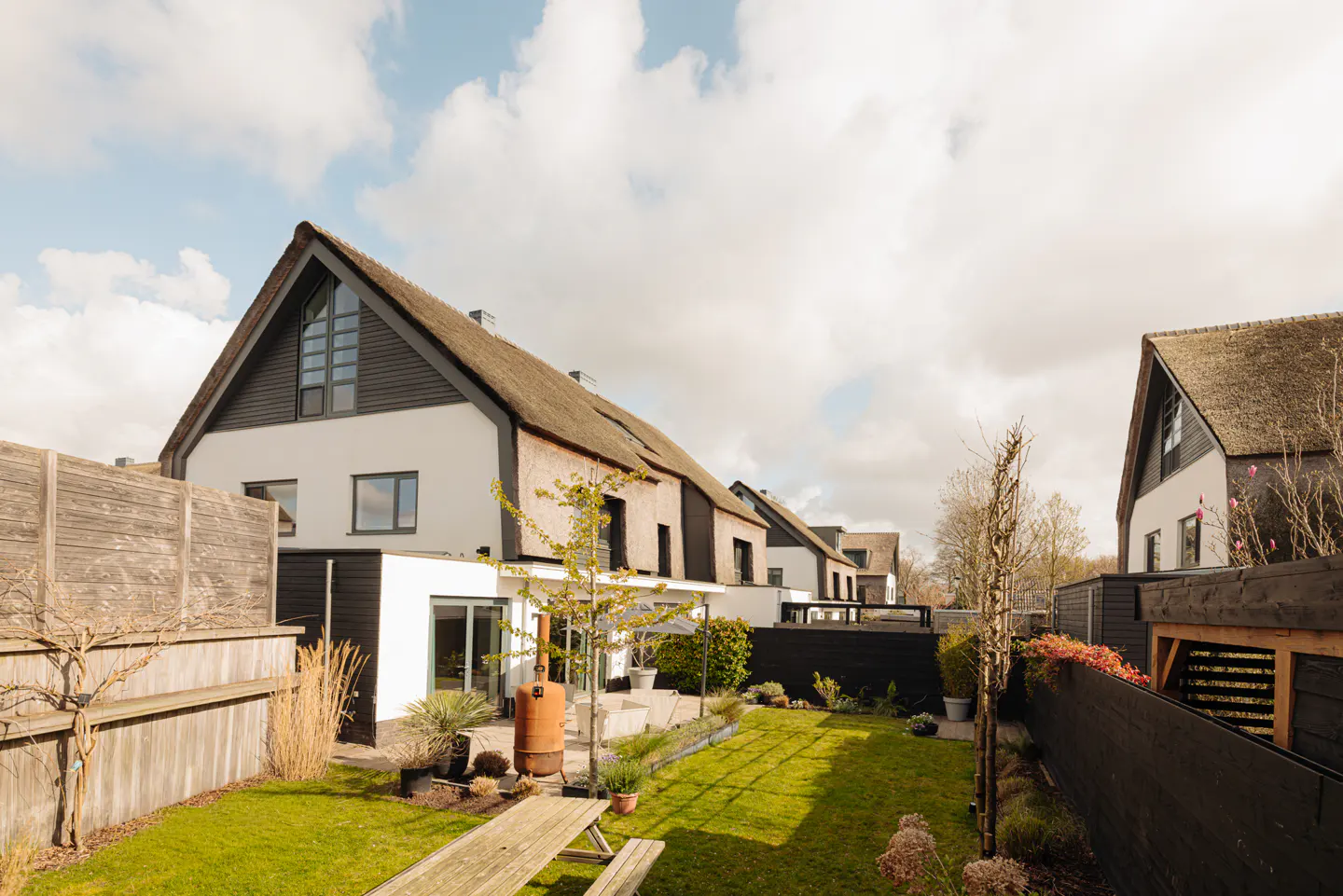 A backyard with a green lawn, a wooden picnic table, and a row of white houses with thatched roofs under a cloudy sky.