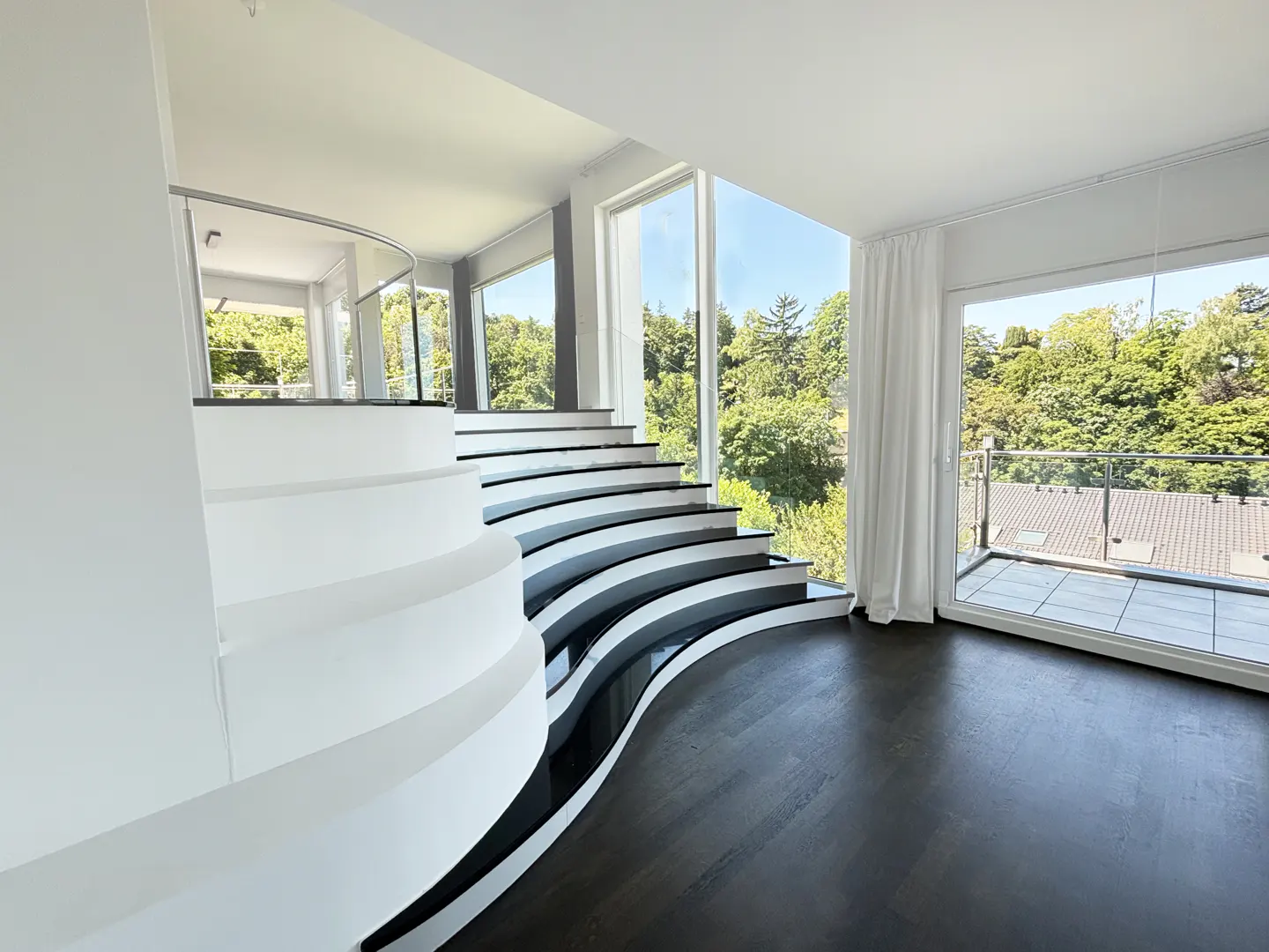 Bright, modern interior with curved white stairs and black treads. Large windows overlook lush green trees and a balcony.