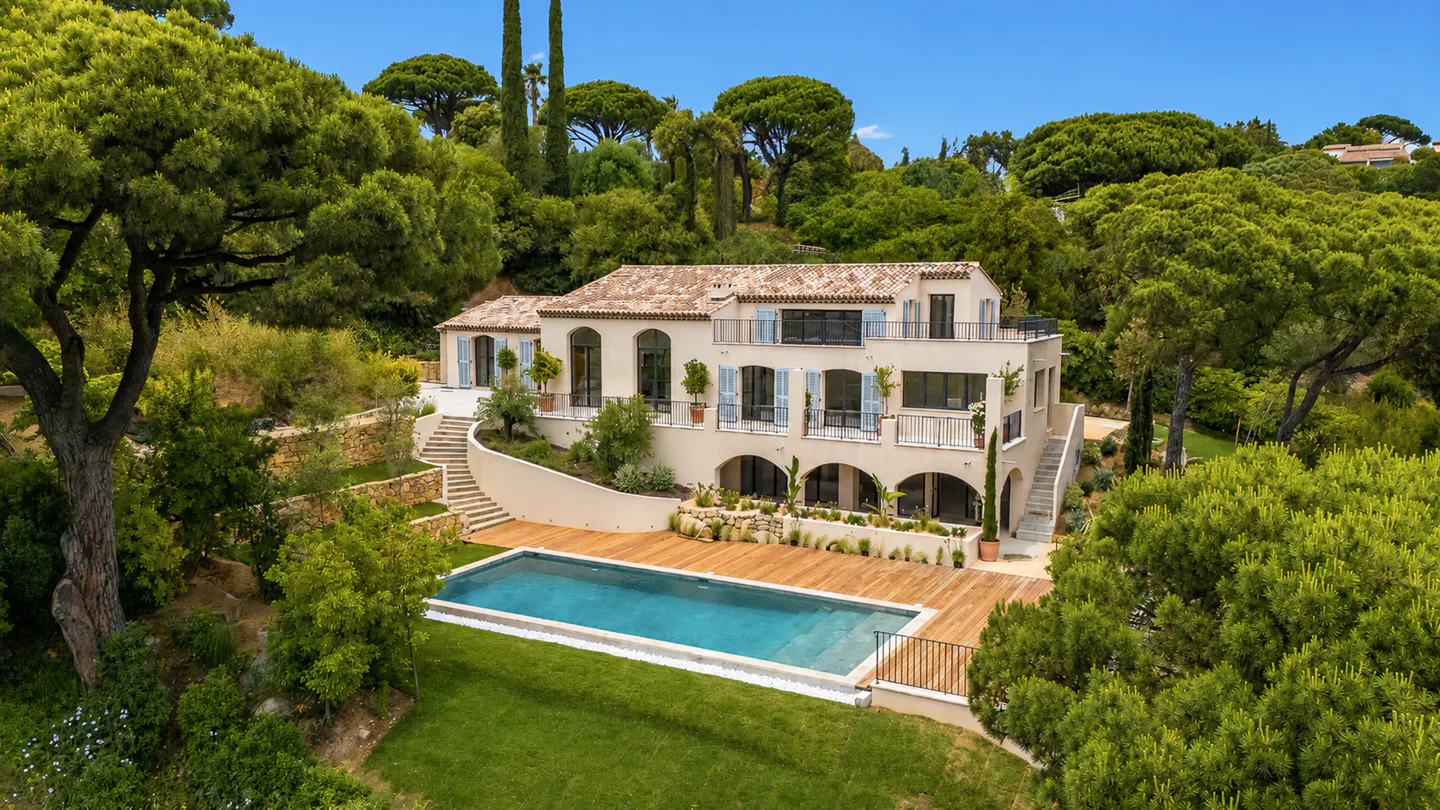 Luxury villa with a pool. The house is beige with blue shutters and a red tile roof, surrounded by green trees.
