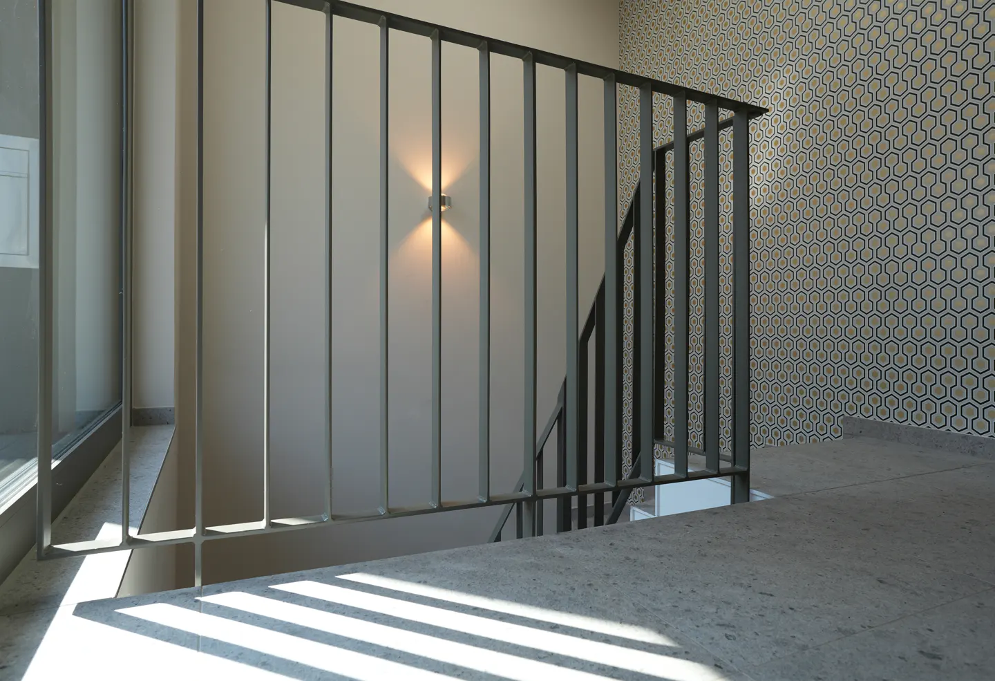 Interior view of a staircase with gray metal railings, patterned wallpaper, and sunlight streaming through a window.