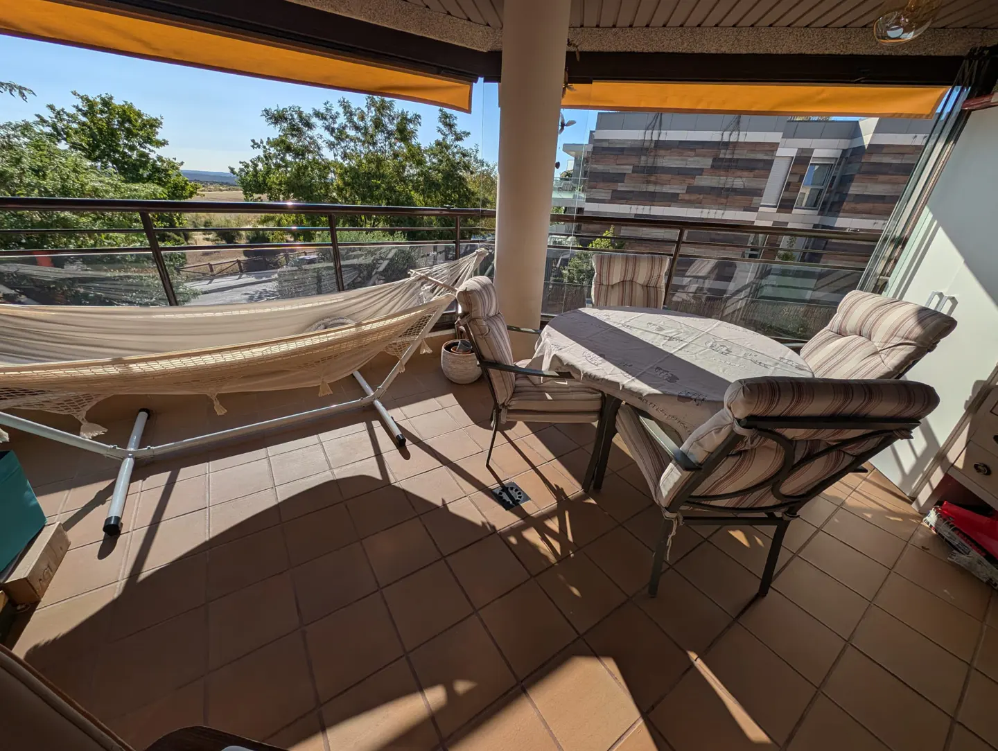 Tiled balcony with a hammock, a round table, and four striped chairs, overlooking trees and a modern building.