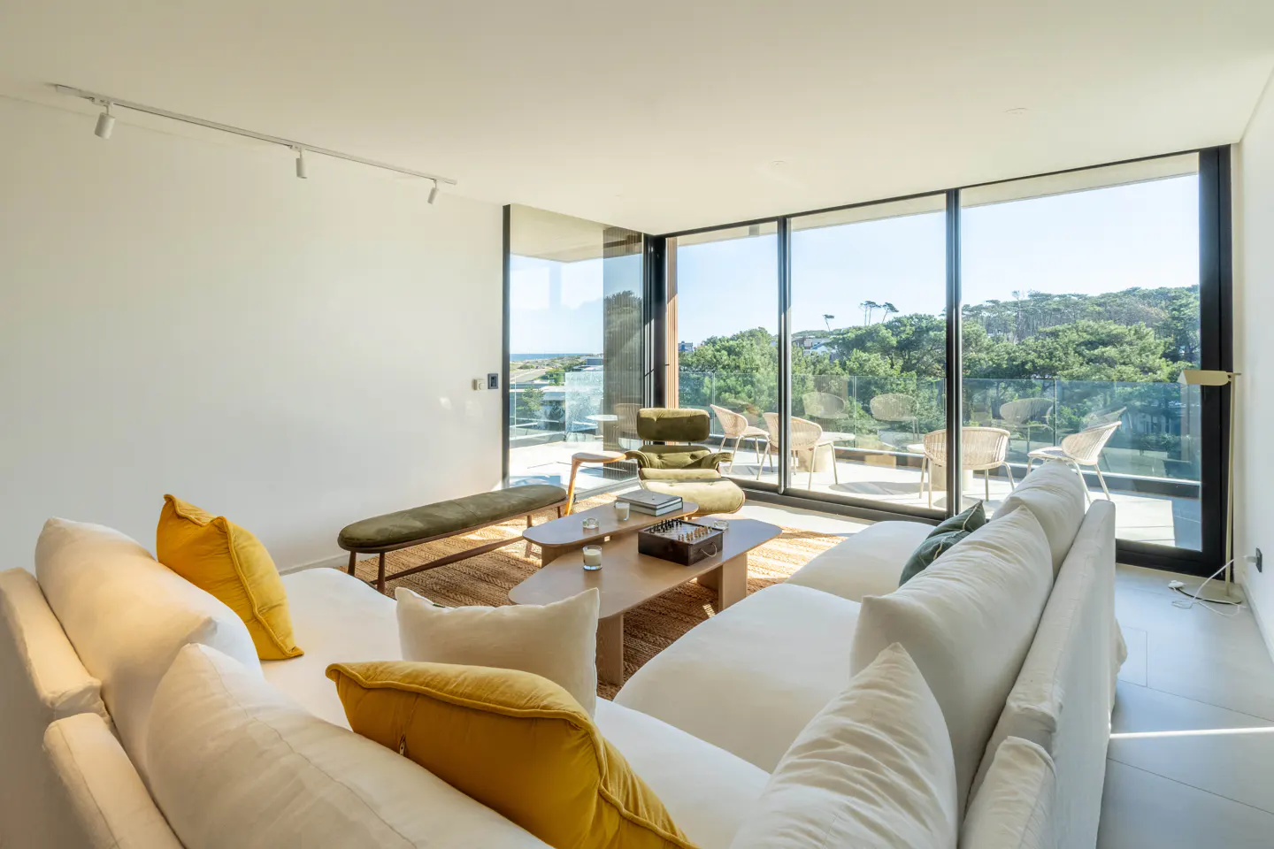 Bright living room with white sofa, yellow pillows, wood coffee table, and floor-to-ceiling windows with a view of trees and ocean.