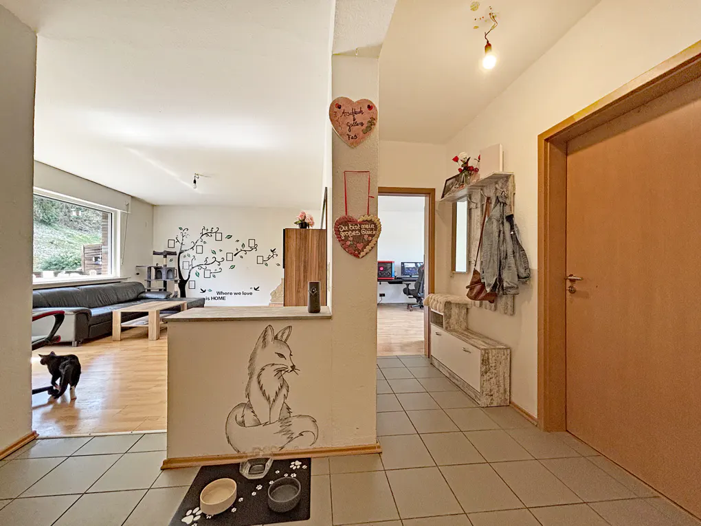 A home's interior view shows a tiled hallway leading to a living room with a black sofa and a wall decal of a tree. A fox drawing is on the wall near pet bowls. A doorway leads to an office space.