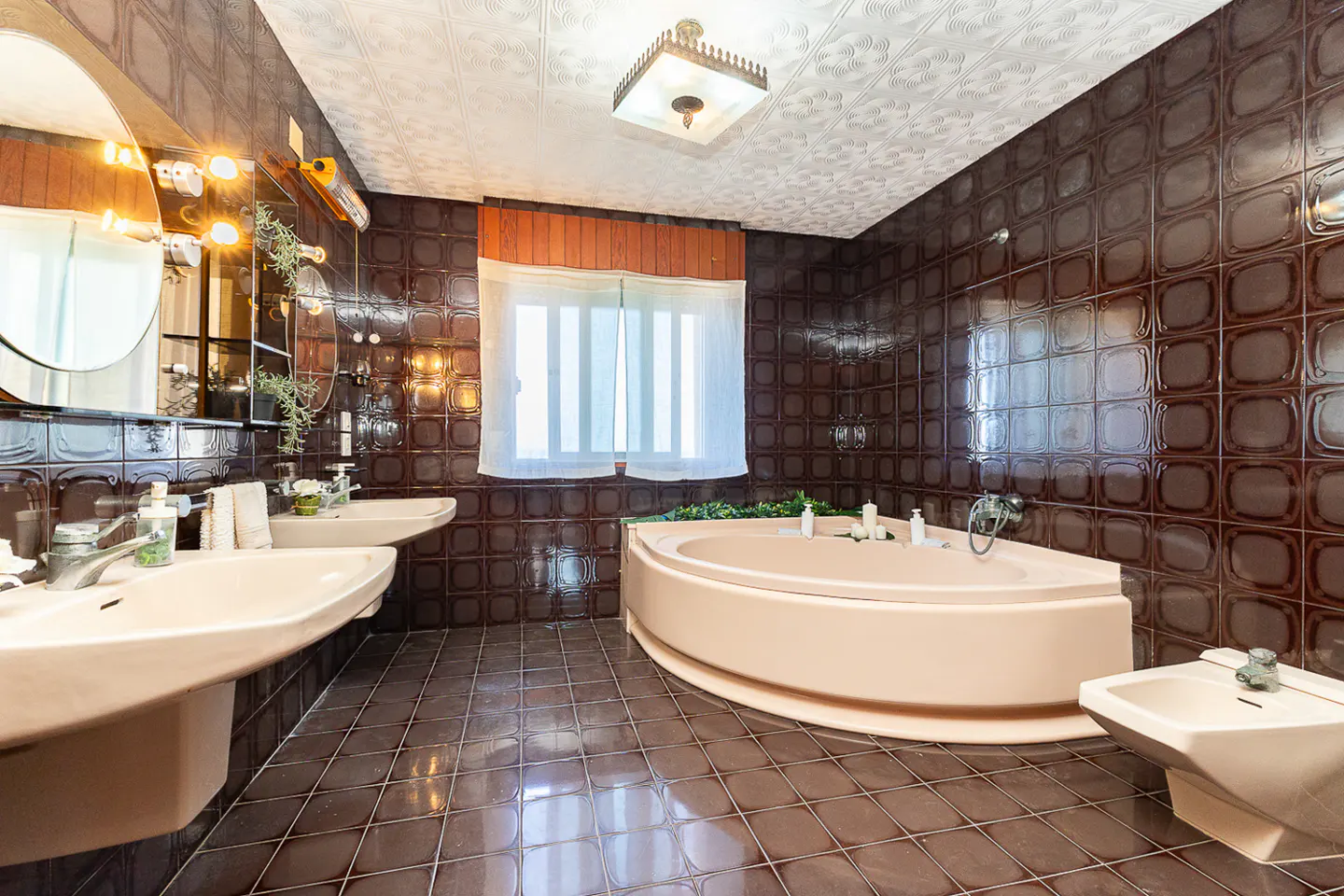 Bathroom with brown square tiles, two sinks, a pink corner tub, and a bidet. A window with white curtains provides natural light.