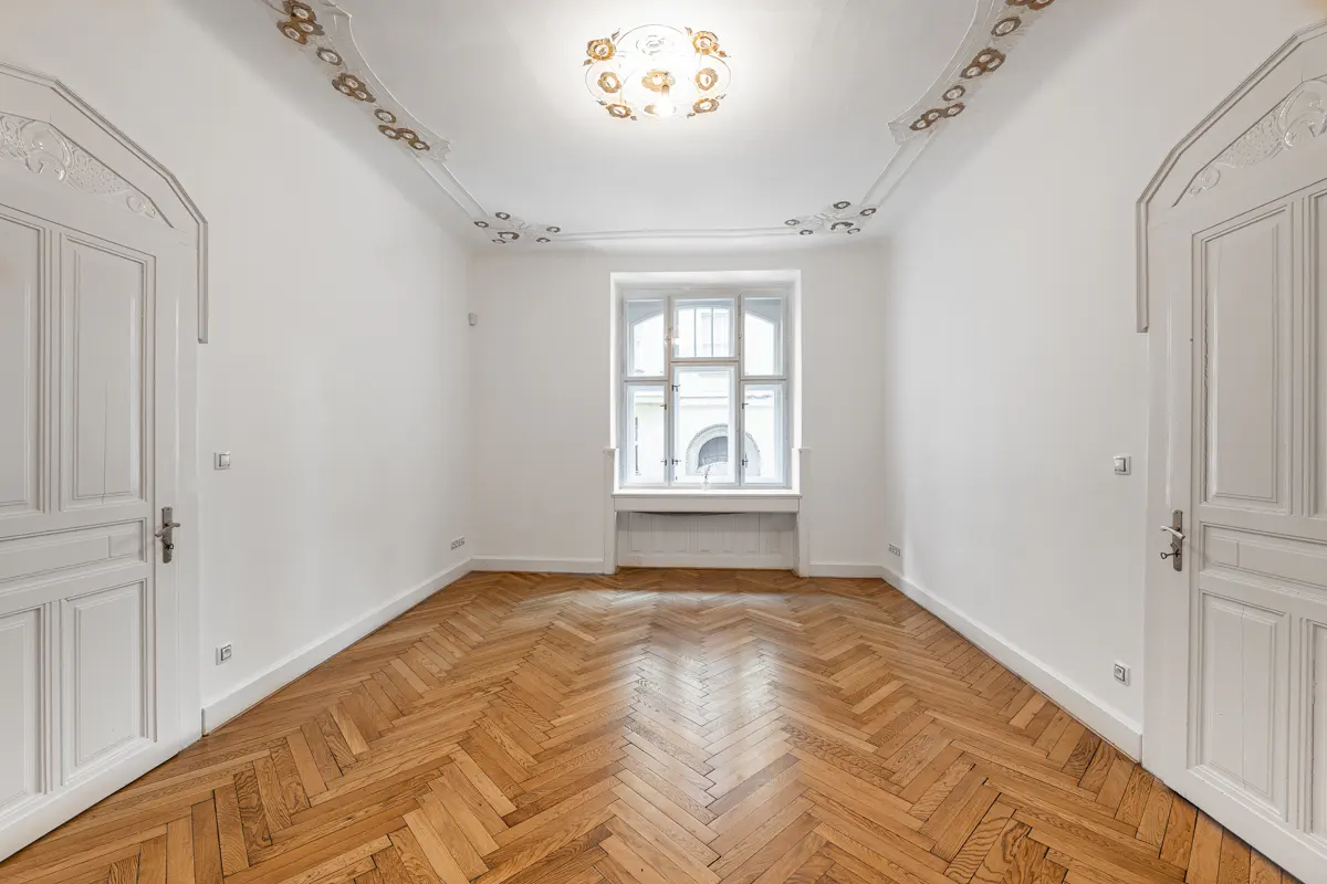 Empty room with herringbone wood floors, white walls, and a window. Two white doors are on either side of the room.