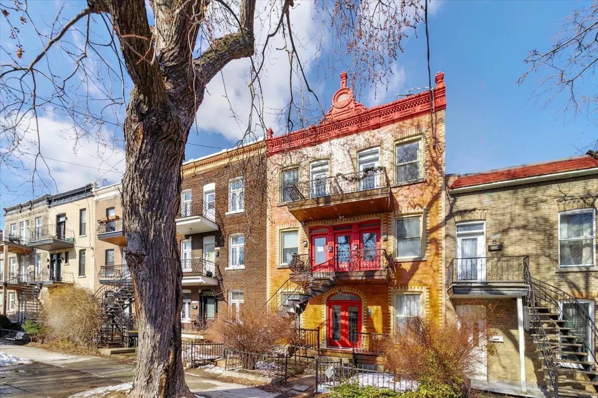 Row of brick townhouses with wrought iron balconies and exterior staircases on a sunny day. A large tree is in the foreground.