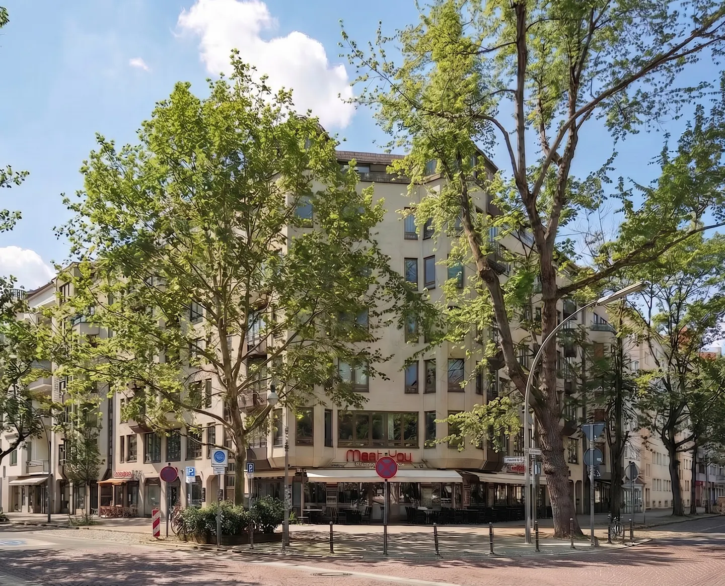 Street view of a light beige building with a "Meet You" restaurant on the ground floor, framed by green trees under a blue sky.