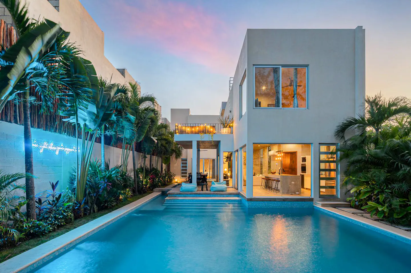 Modern white house with a blue pool, palm trees, and a neon sign on the wall. The sky is pink and blue.