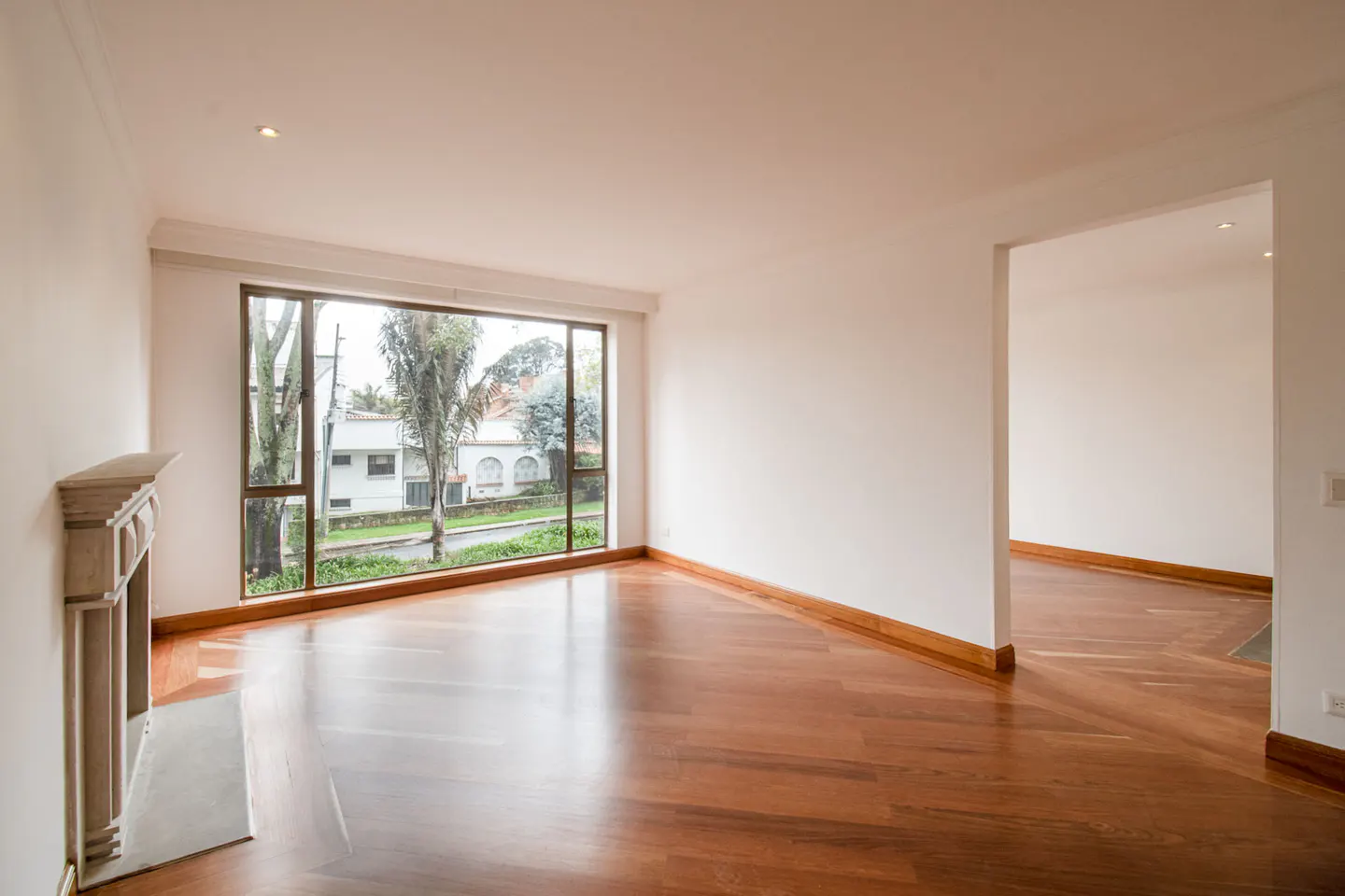 Bright, empty room with hardwood floors, white walls, and a large window showing a residential street. A decorative fireplace is on the left.