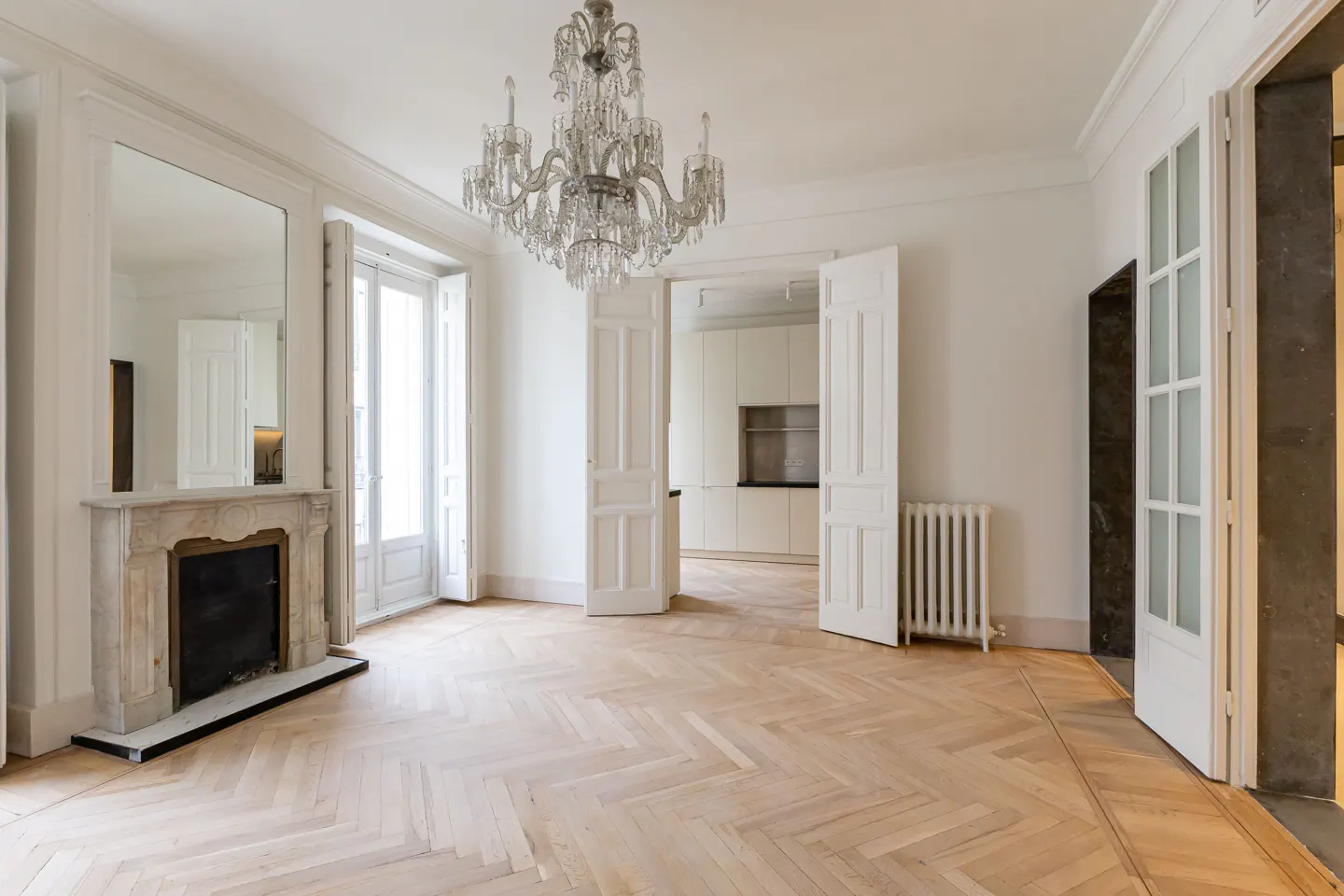 Bright, empty room with herringbone wood floors, white walls, and a crystal chandelier. A marble fireplace and open doorways add architectural detail.