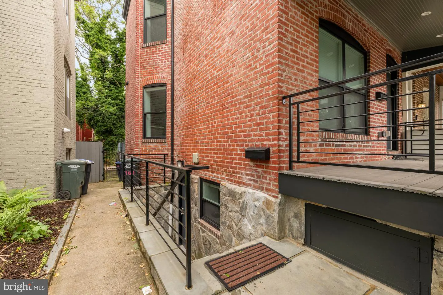 Exterior view of a red brick house with black window frames and railings, next to a gray building and a narrow walkway.