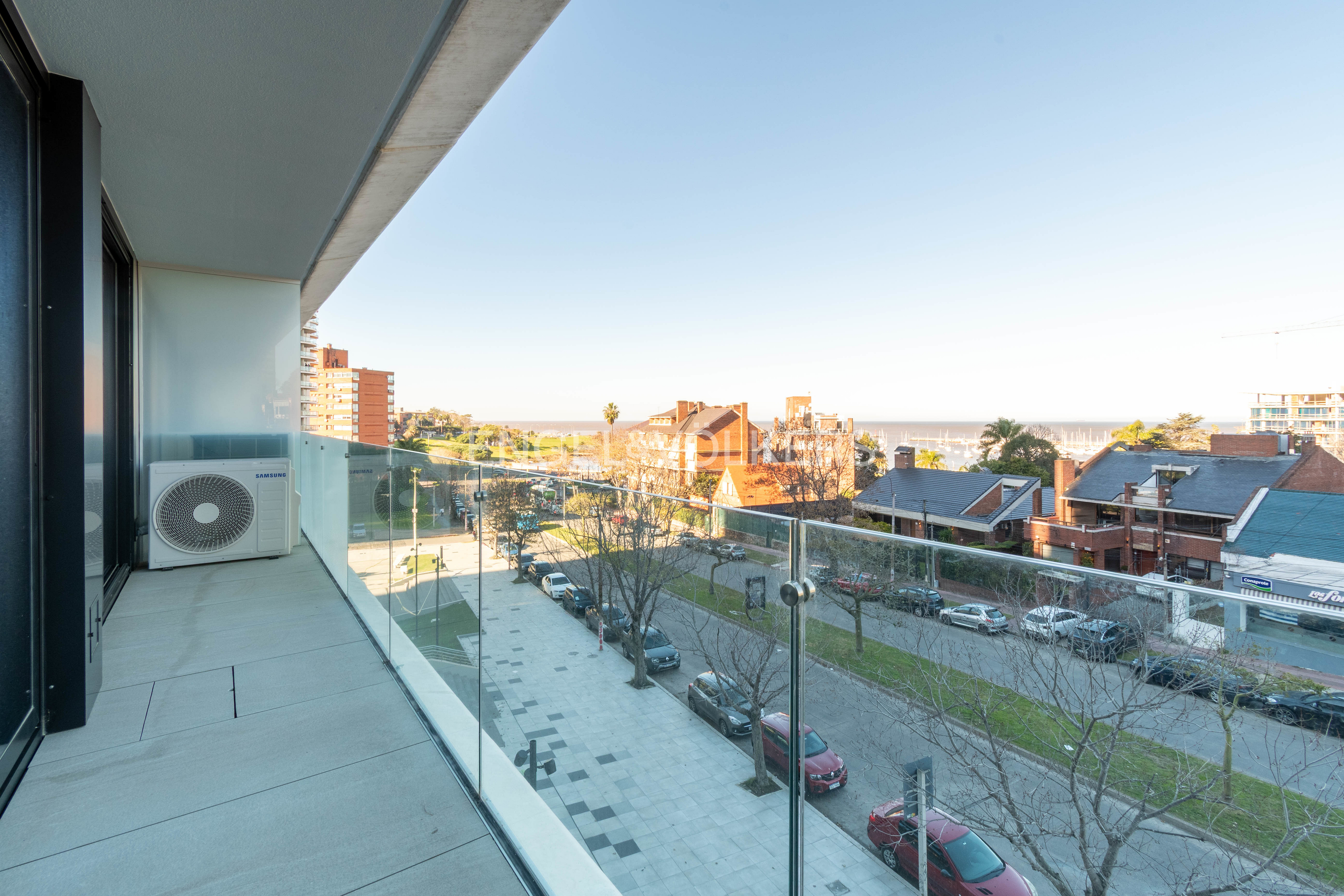 View from a modern balcony with a glass railing overlooking a street with parked cars, trees, and distant coastal buildings.