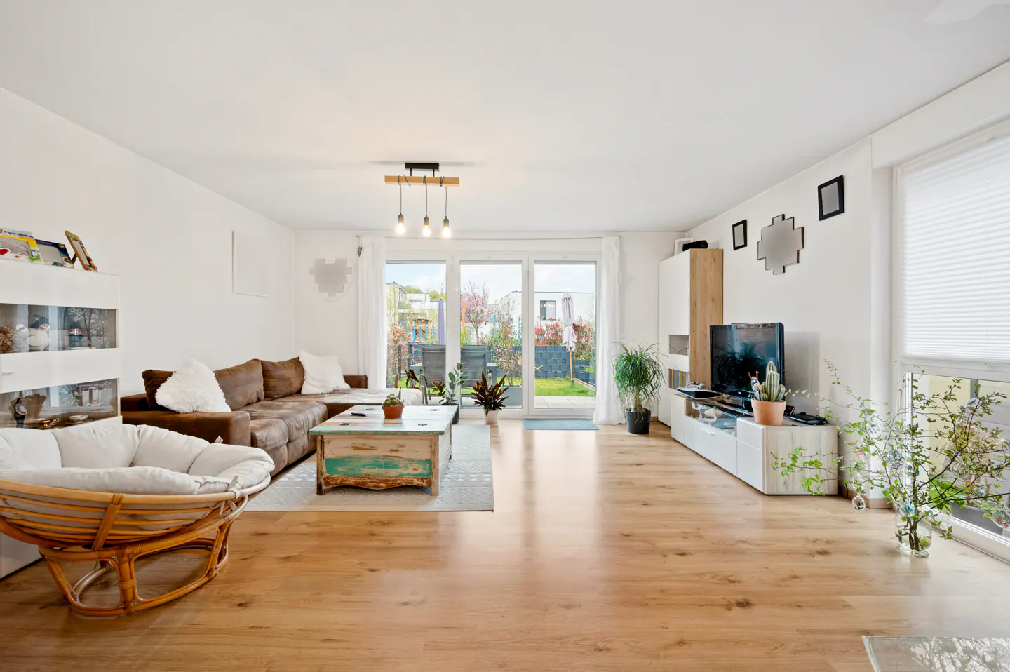 Bright living room with wood floors, brown sectional sofa, rattan chair, and sliding glass doors to a garden.