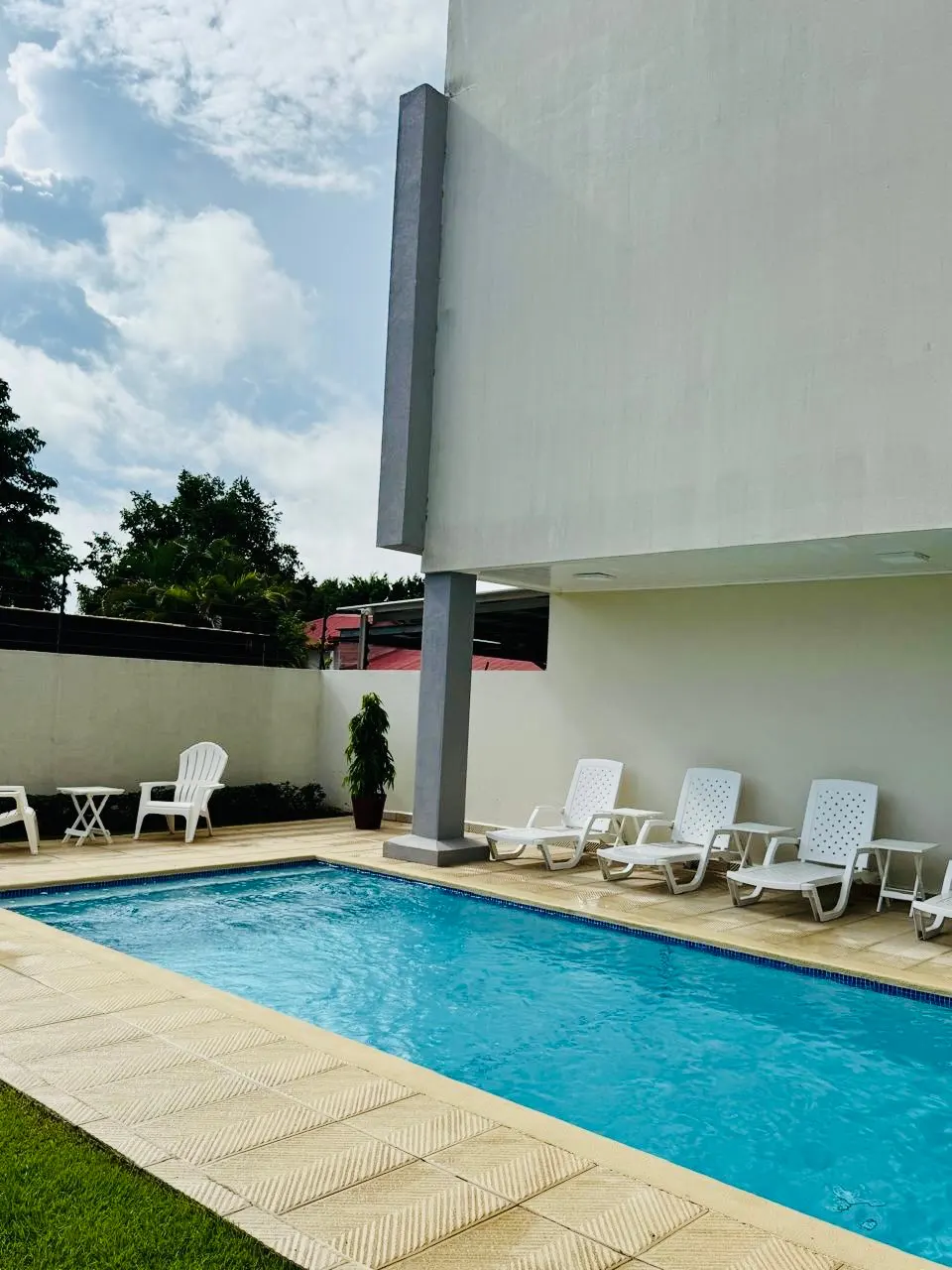 Outdoor pool with lounge chairs and a modern building. Blue water, beige tiles, and a cloudy sky.