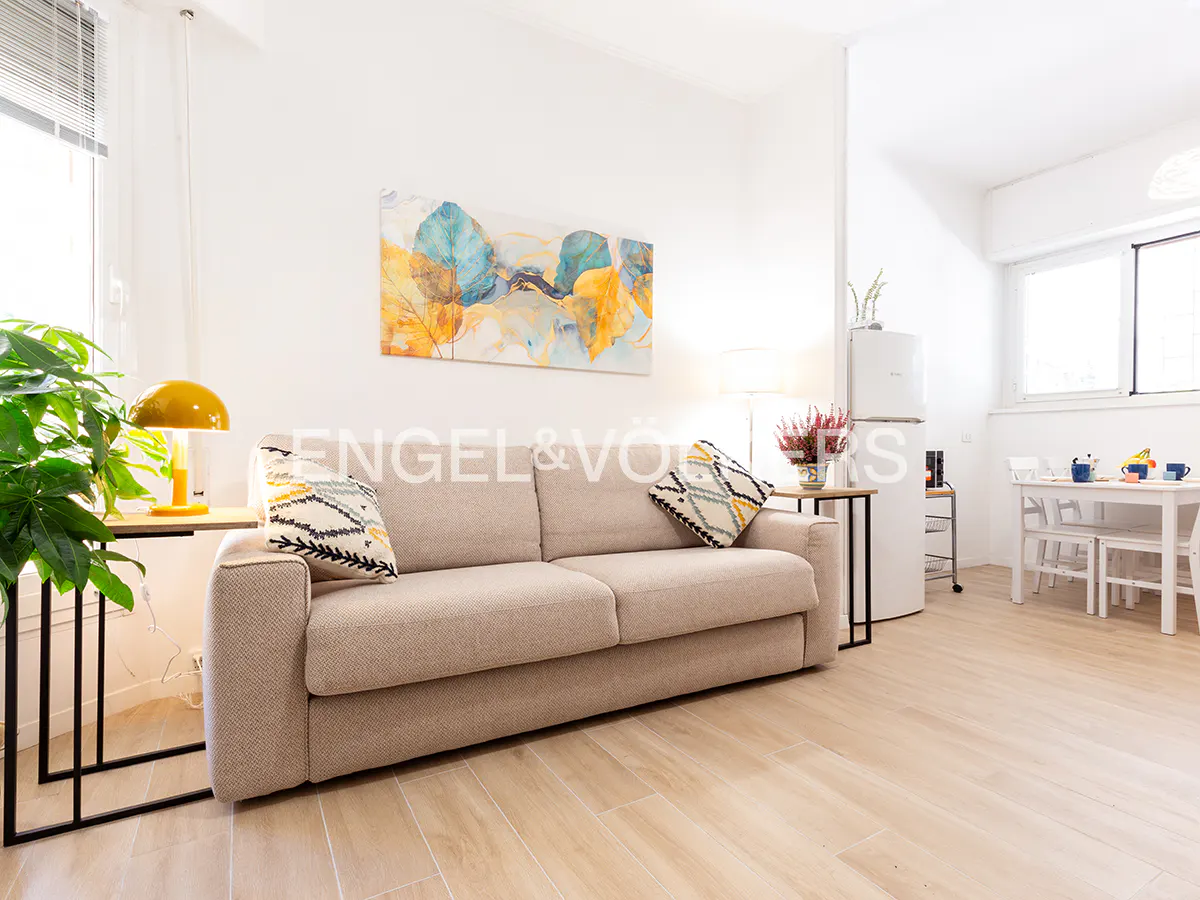 Bright apartment interior with a beige sofa, patterned pillows, and a colorful leaf painting above. A white fridge and dining table are visible in the background.