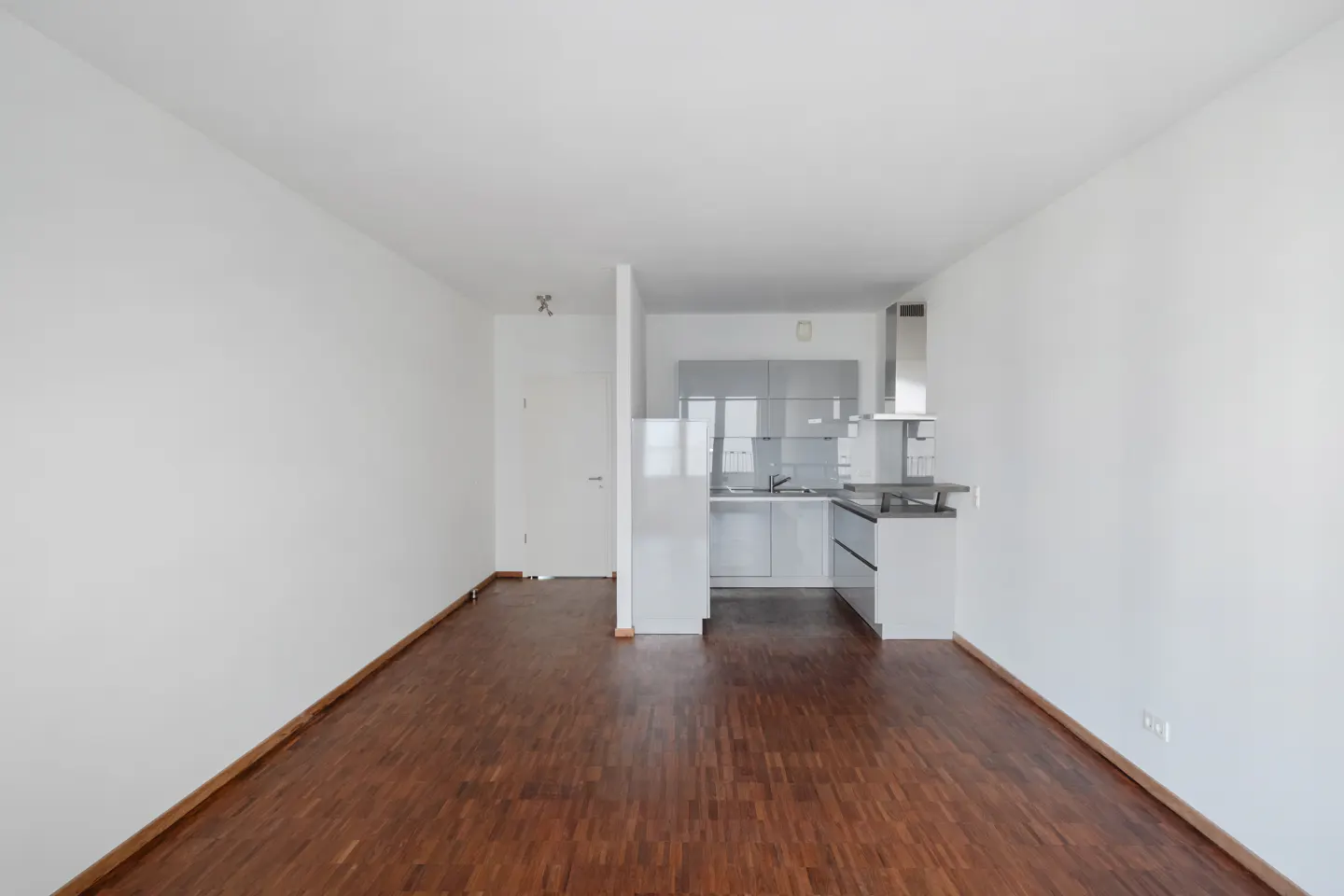 An empty room with hardwood floors and white walls, leading to a modern kitchen with gray cabinets.